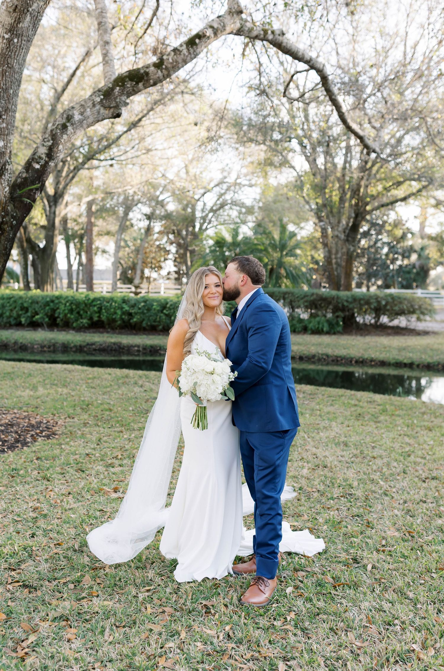 A bride in a white gown and groom in a navy suit share a romantic moment outdoors under oak trees.