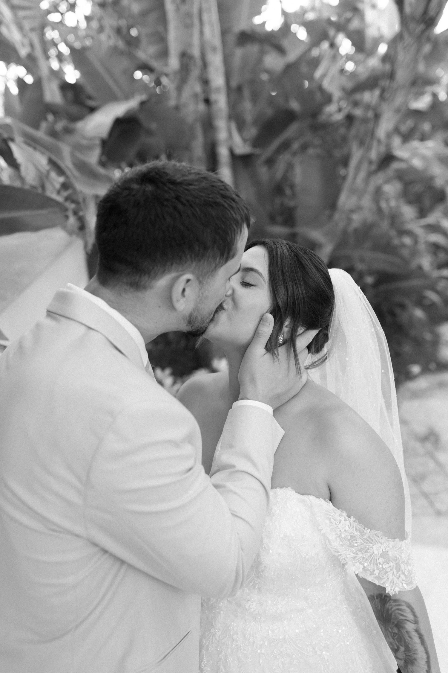 Black and white photo of groom tenderly cupping bride's face during a passionate wedding kiss outdoors.