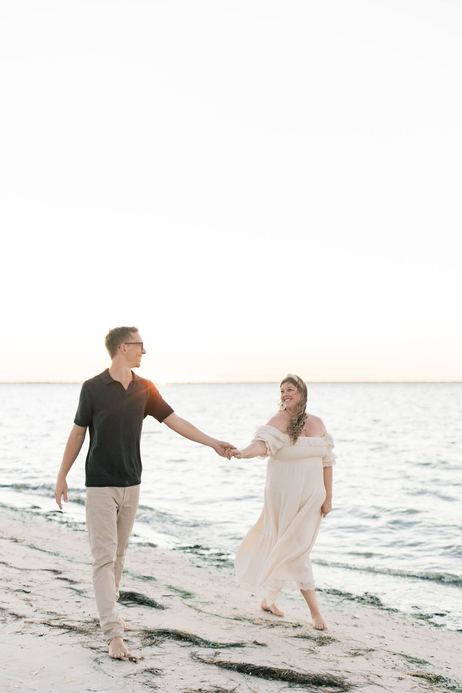Couple holding hands walking barefoot on a sandy beach at sunset, woman in white dress, man in black shirt.