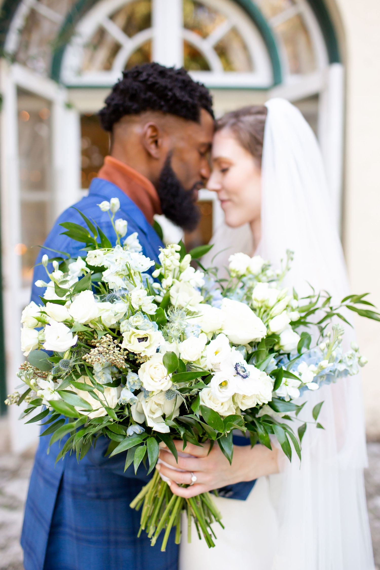 Interracial couple sharing an intimate moment at their wedding, with a lush white and green bridal bouquet in the foreground.