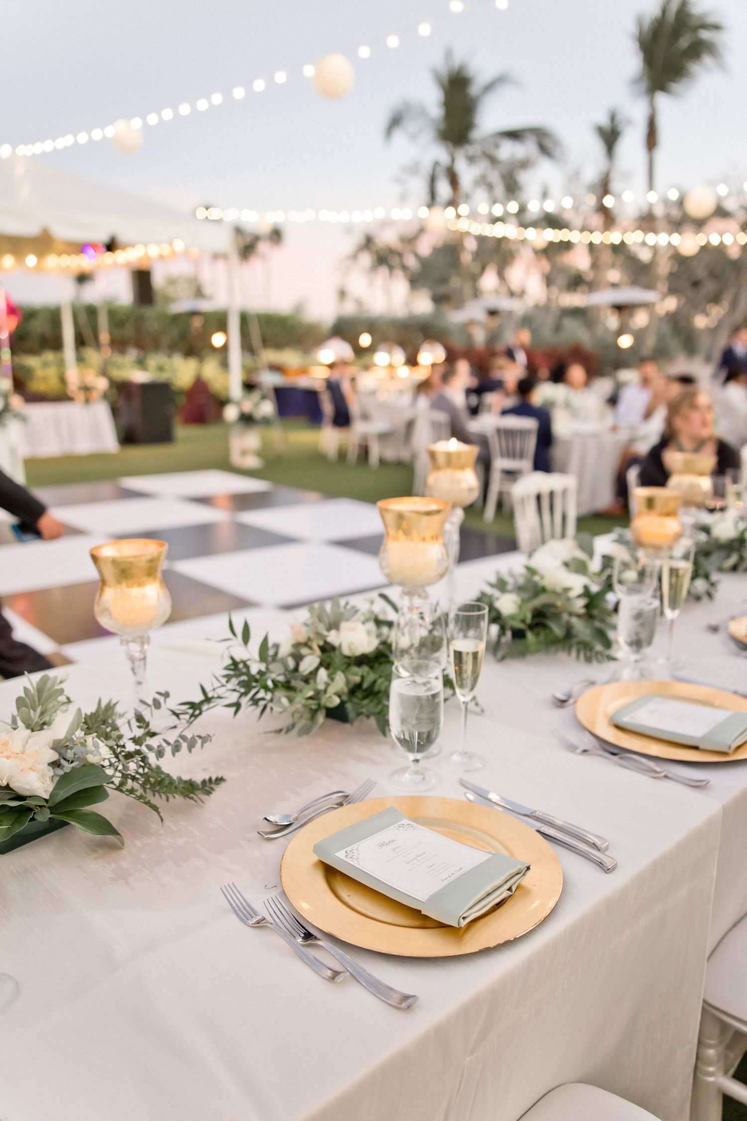 Elegant wedding reception tables with gold chargers and greenery centerpieces under string lights.