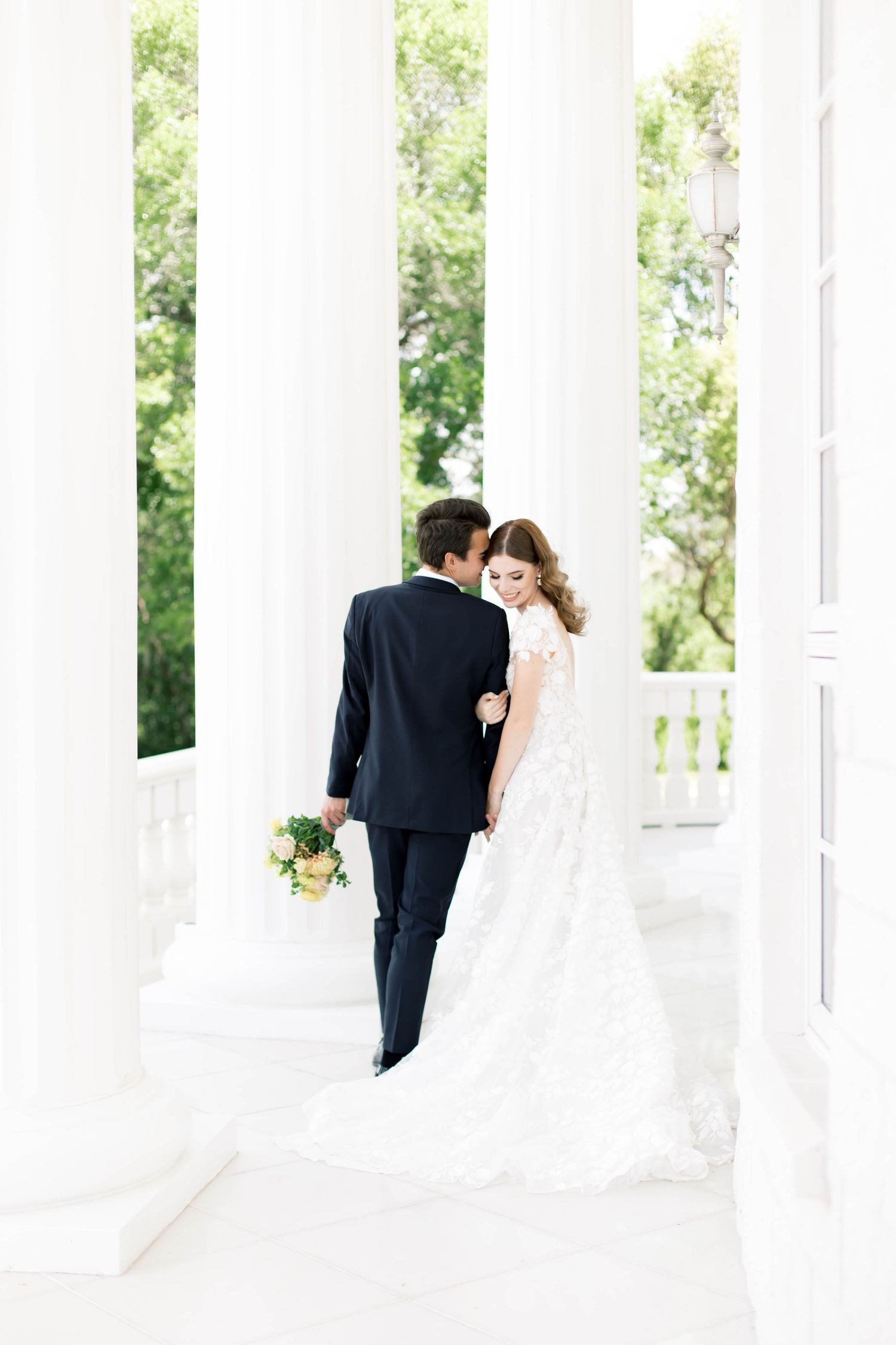 Bride and groom stand romantically among white columns, she in a lace gown holding a bouquet, he in a dark suit.