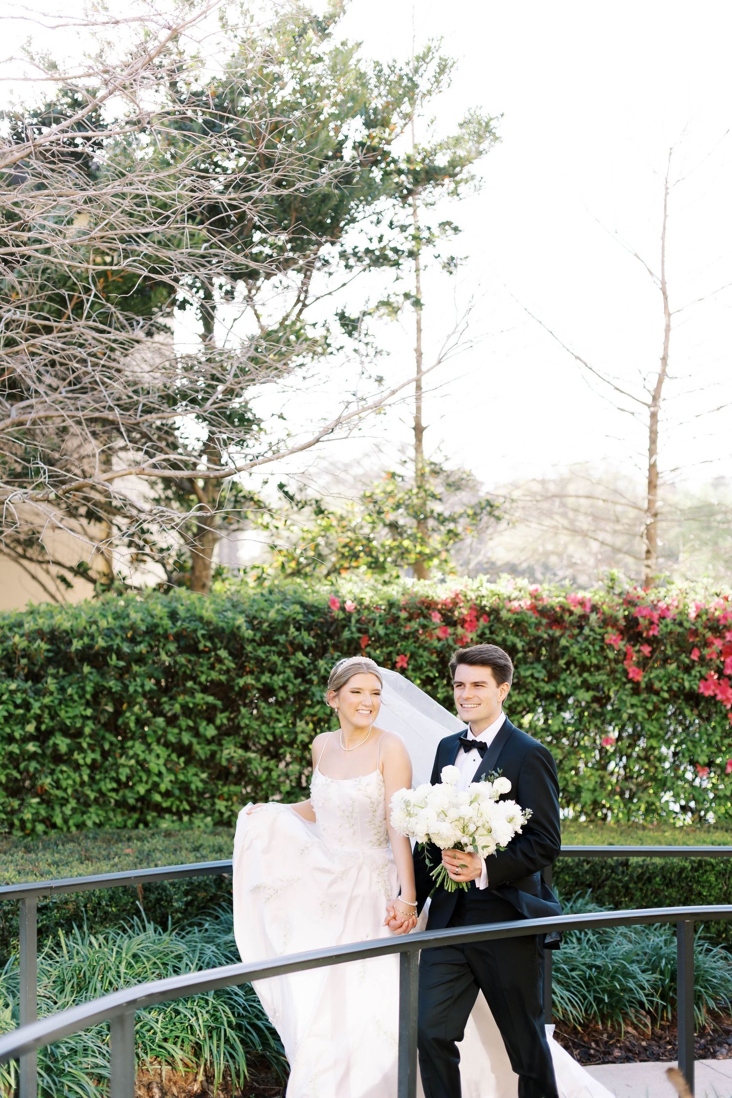 Bride and groom hold hands on a curved bridge surrounded by lush greenery and red flowers on their wedding day.