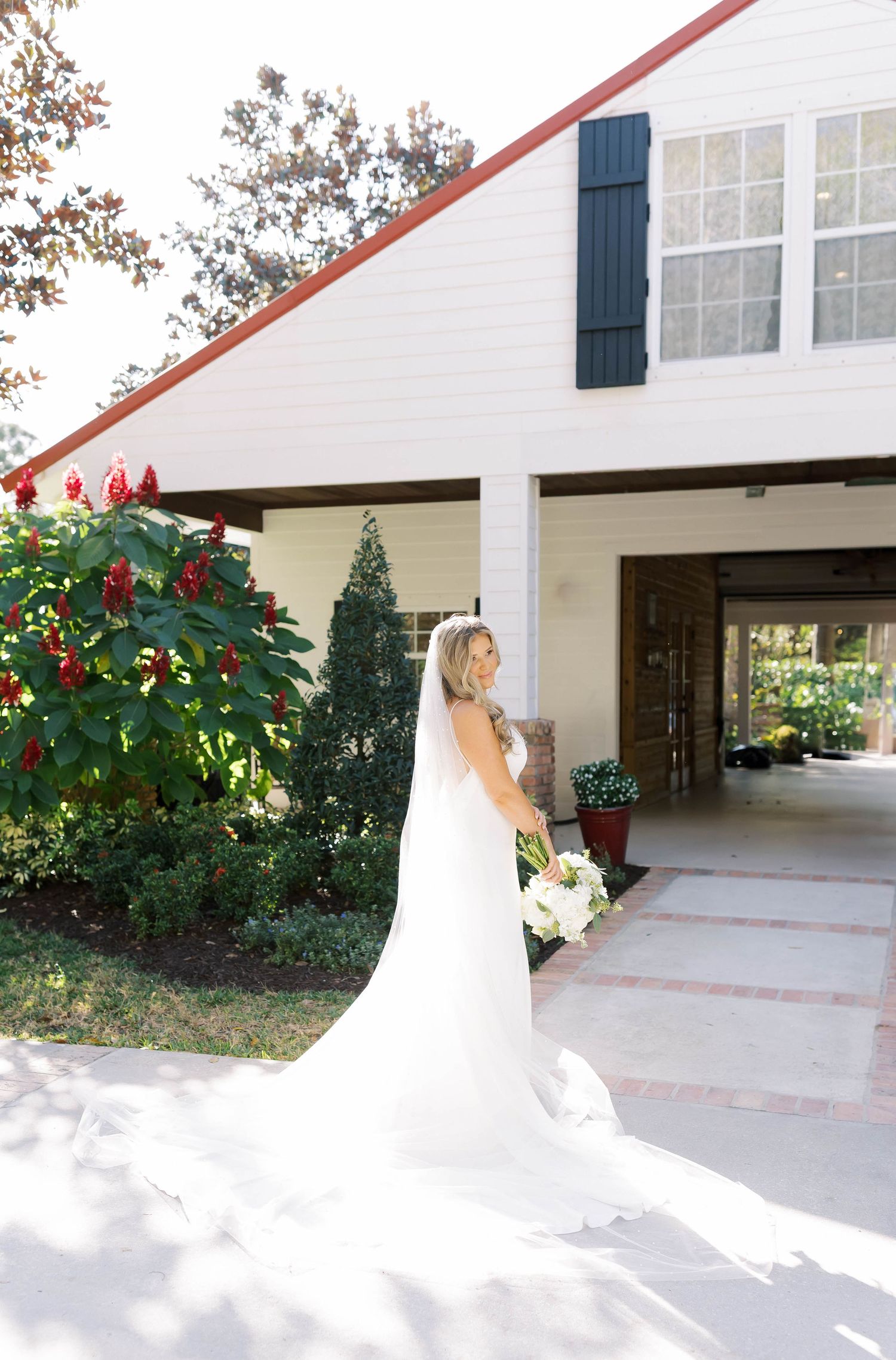 Bride in flowing white gown poses in front of classic white venue with navy shutters.