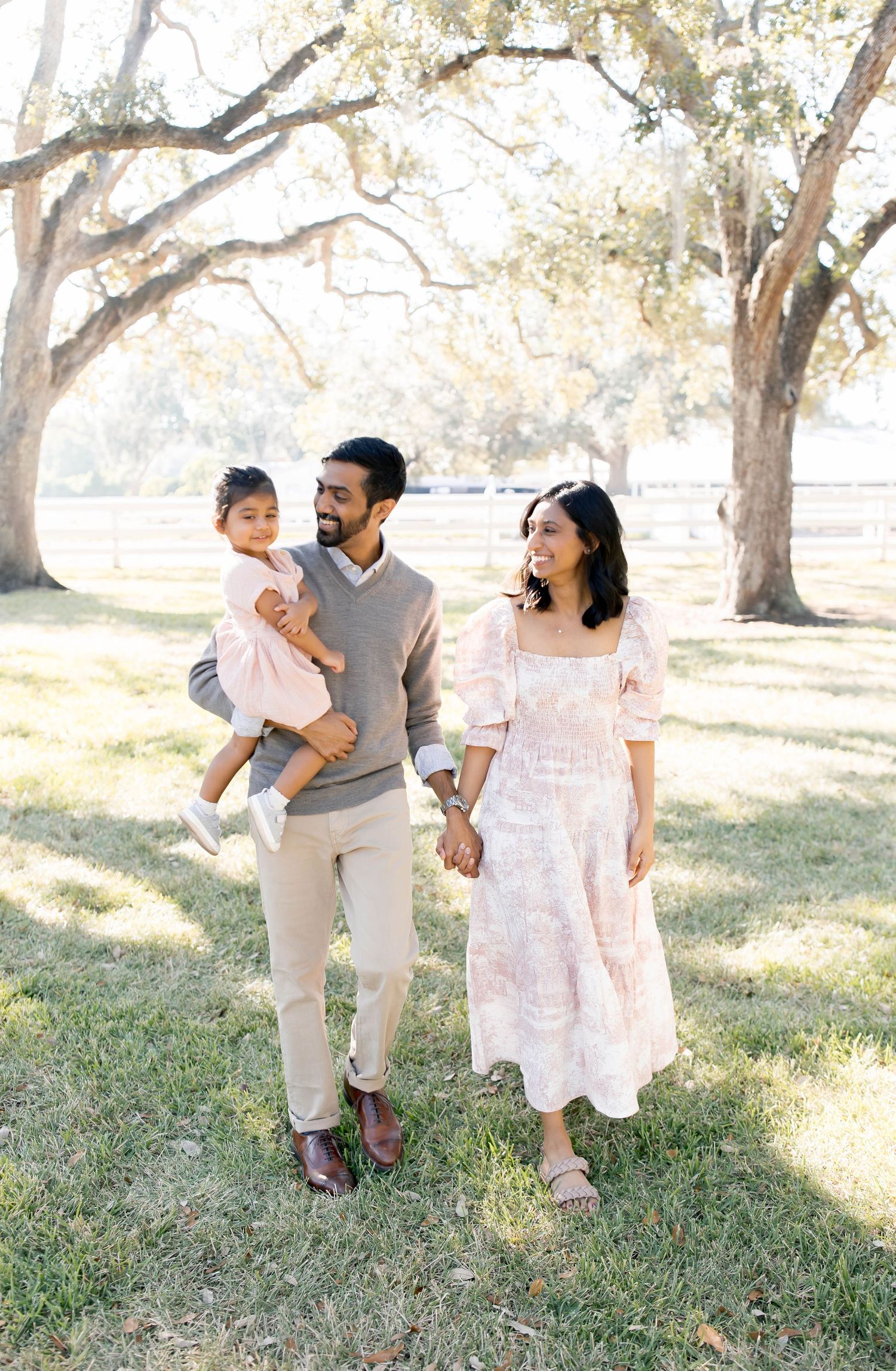 Family walking together through a sunlit grove of oak trees in coordinated pastel outfits.