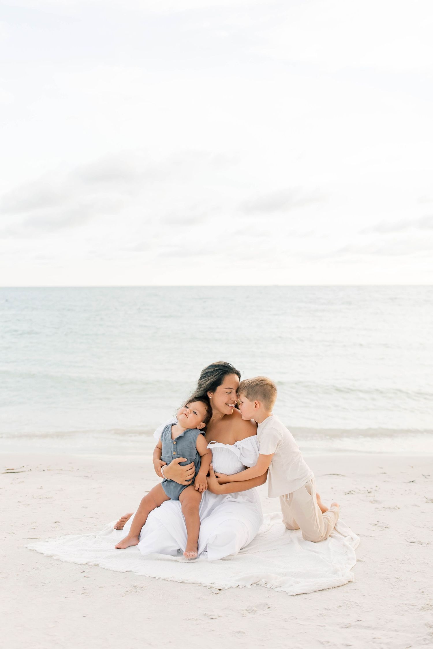 A family enjoys a playful moment lying together on a white blanket at the beach.