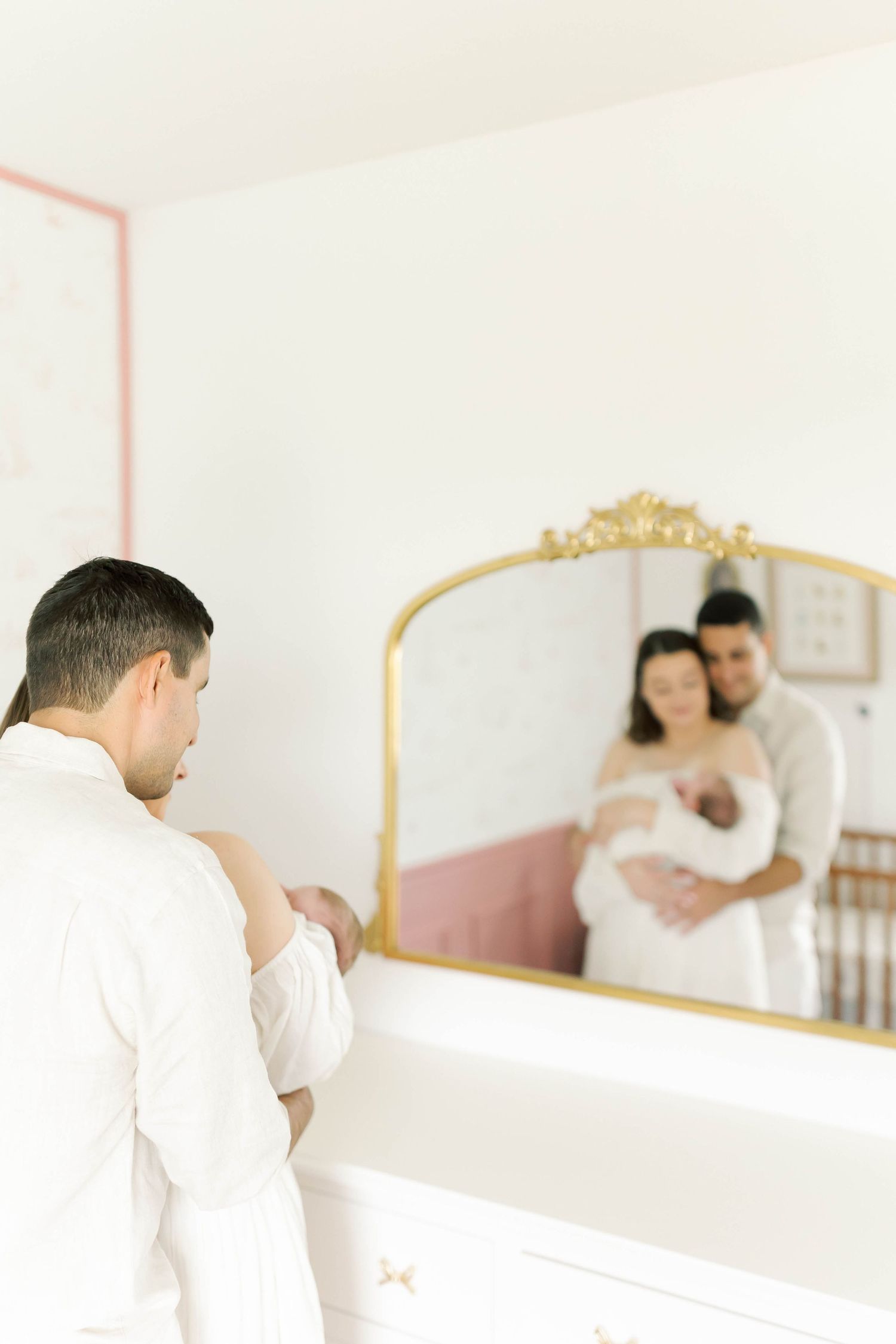 A reflection in an ornate gold mirror shows a family moment in a pink and white nursery.