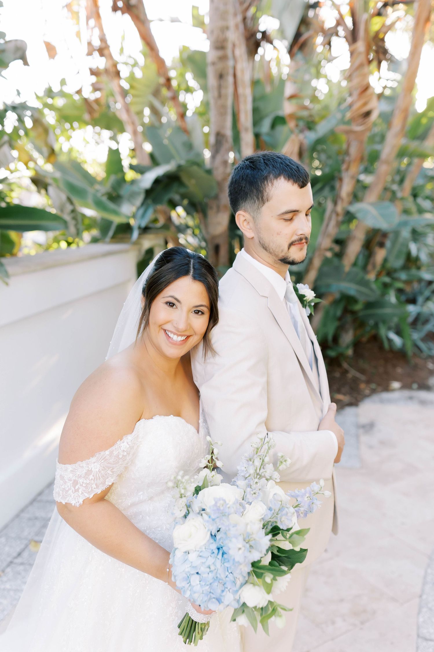 Smiling bride holding blue hydrangea bouquet poses back-to-back with groom in tan suit amid lush greenery.