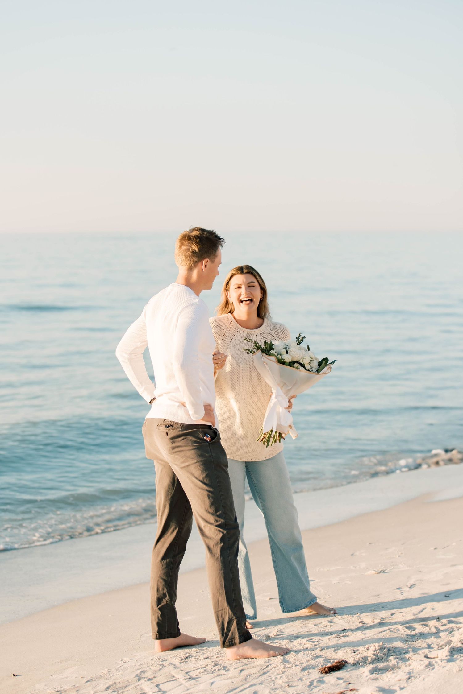 A couple in white and beige clothing stands on a sunny beach holding a bouquet of flowers.
