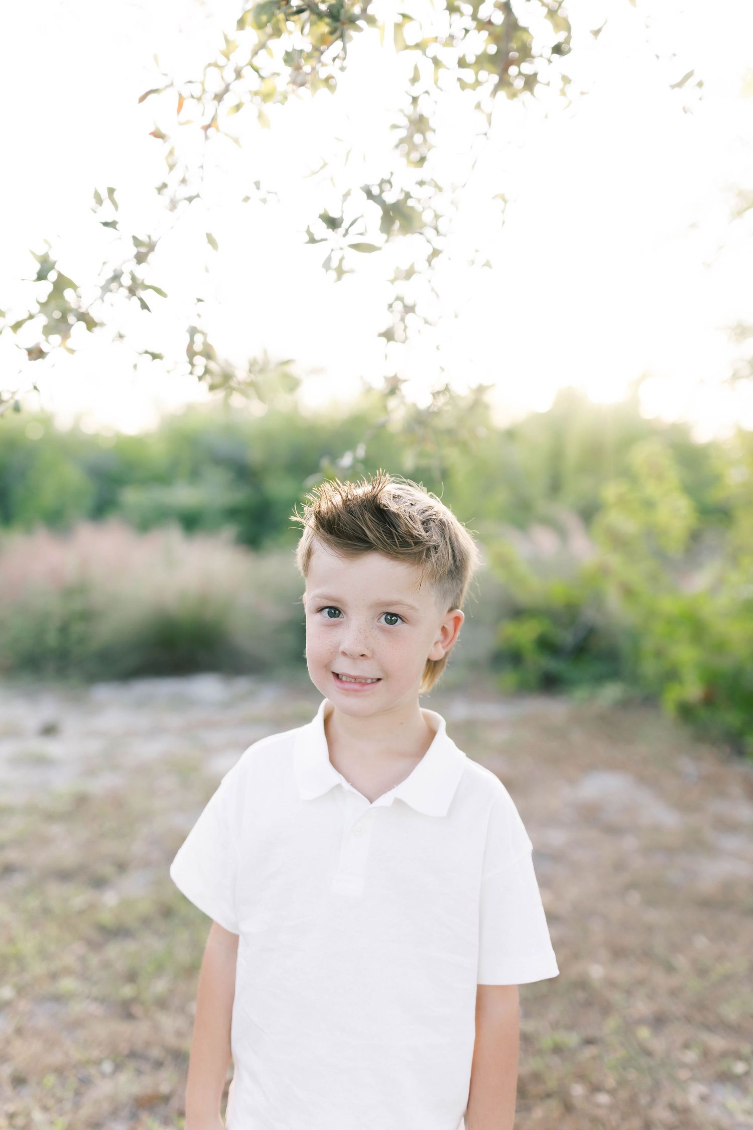 Portrait of a young person in a white polo shirt standing outdoors with sunlit foliage in the background.