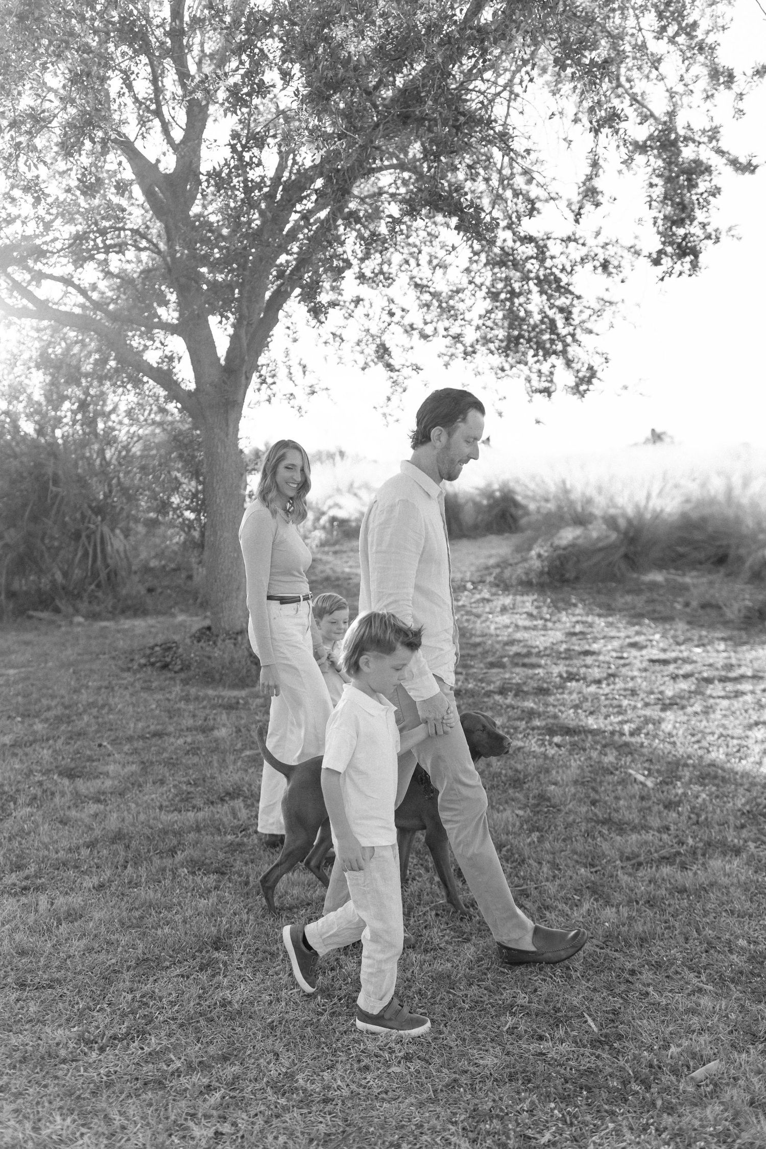 Family walking together on a grassy hillside under a large tree in black and white.