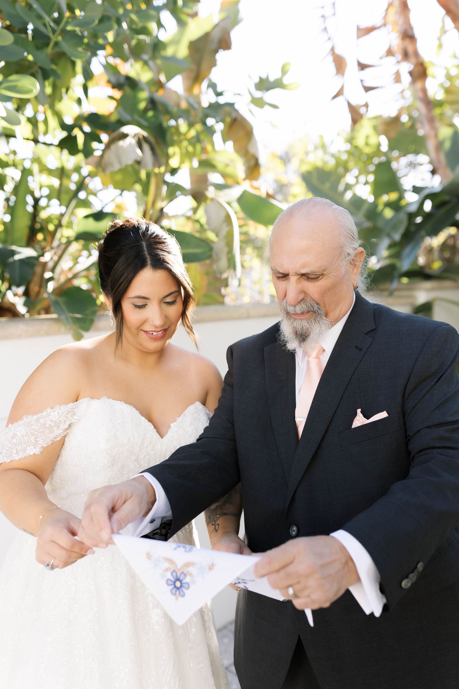 Bride in lace gown and older man in black suit examine a handkerchief together outdoors at a wedding.