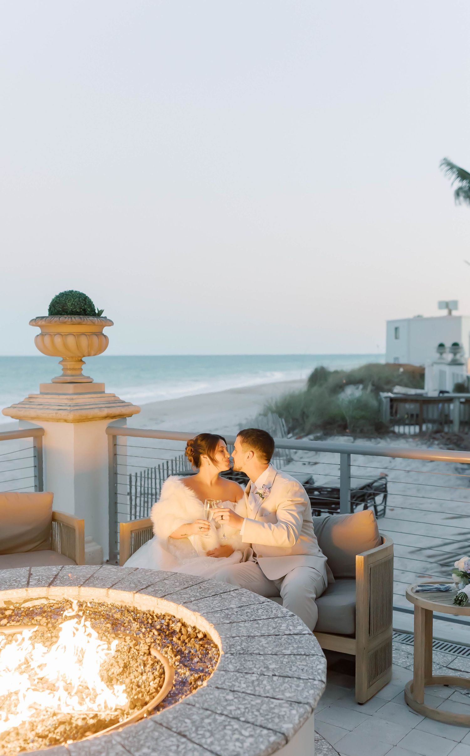 Newlyweds share a kiss beside a glowing fire pit on an oceanfront terrace at twilight, holding champagne glasses.