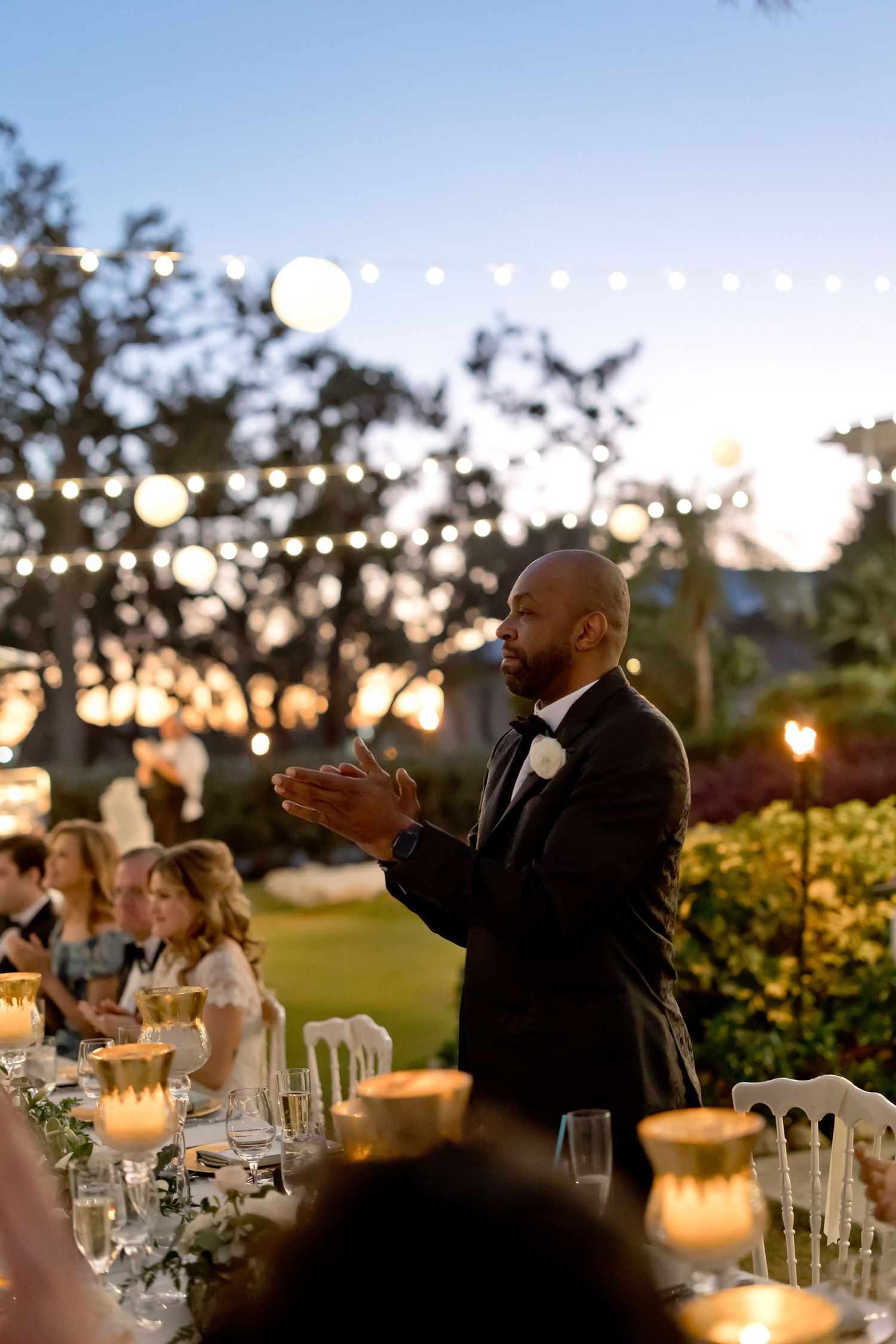 Wedding guests applauding during outdoor reception with string lights and palm trees.