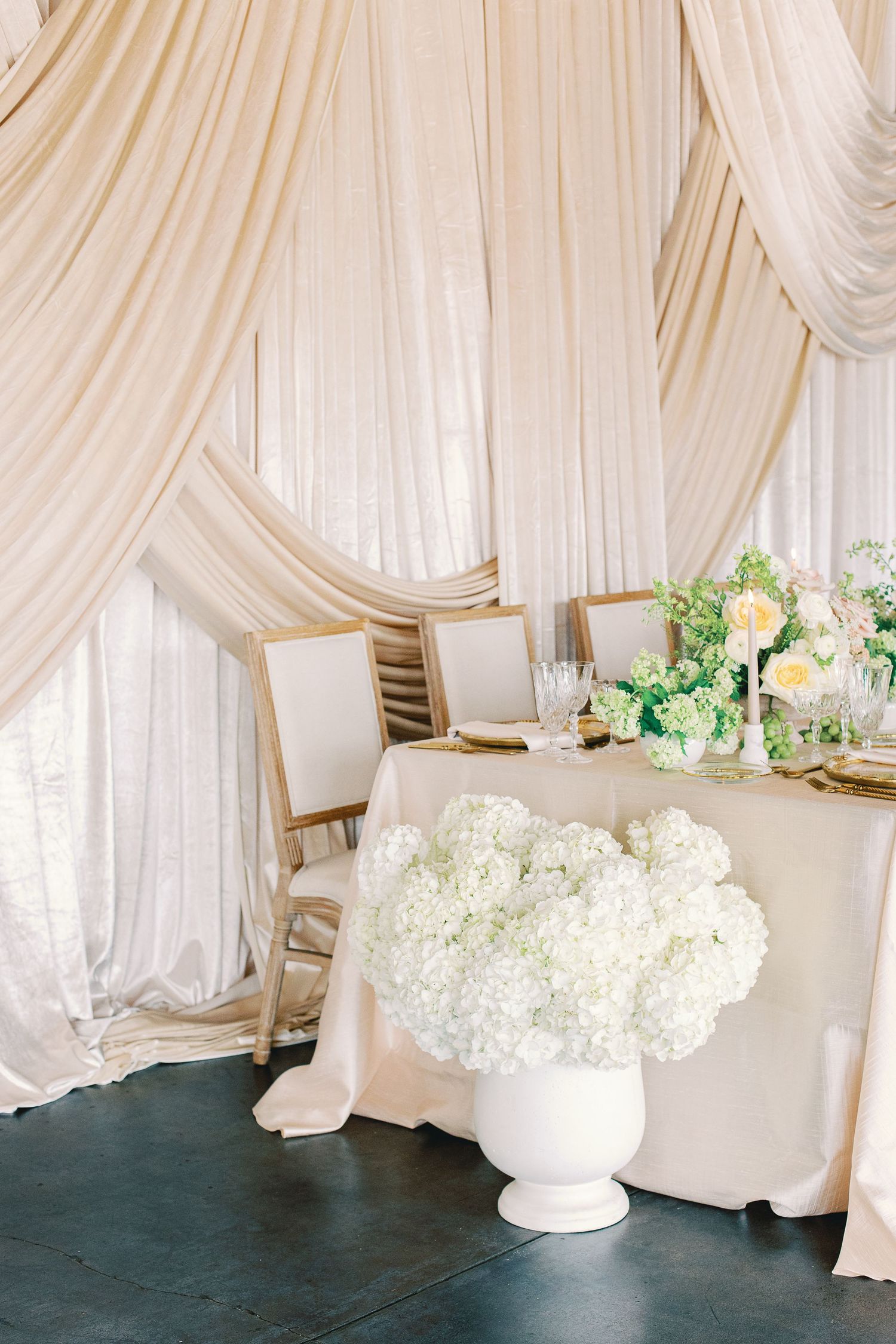 Elegant wedding sweetheart table with white hydrangeas, champagne draping, and gold chairs in a luxurious reception setting.