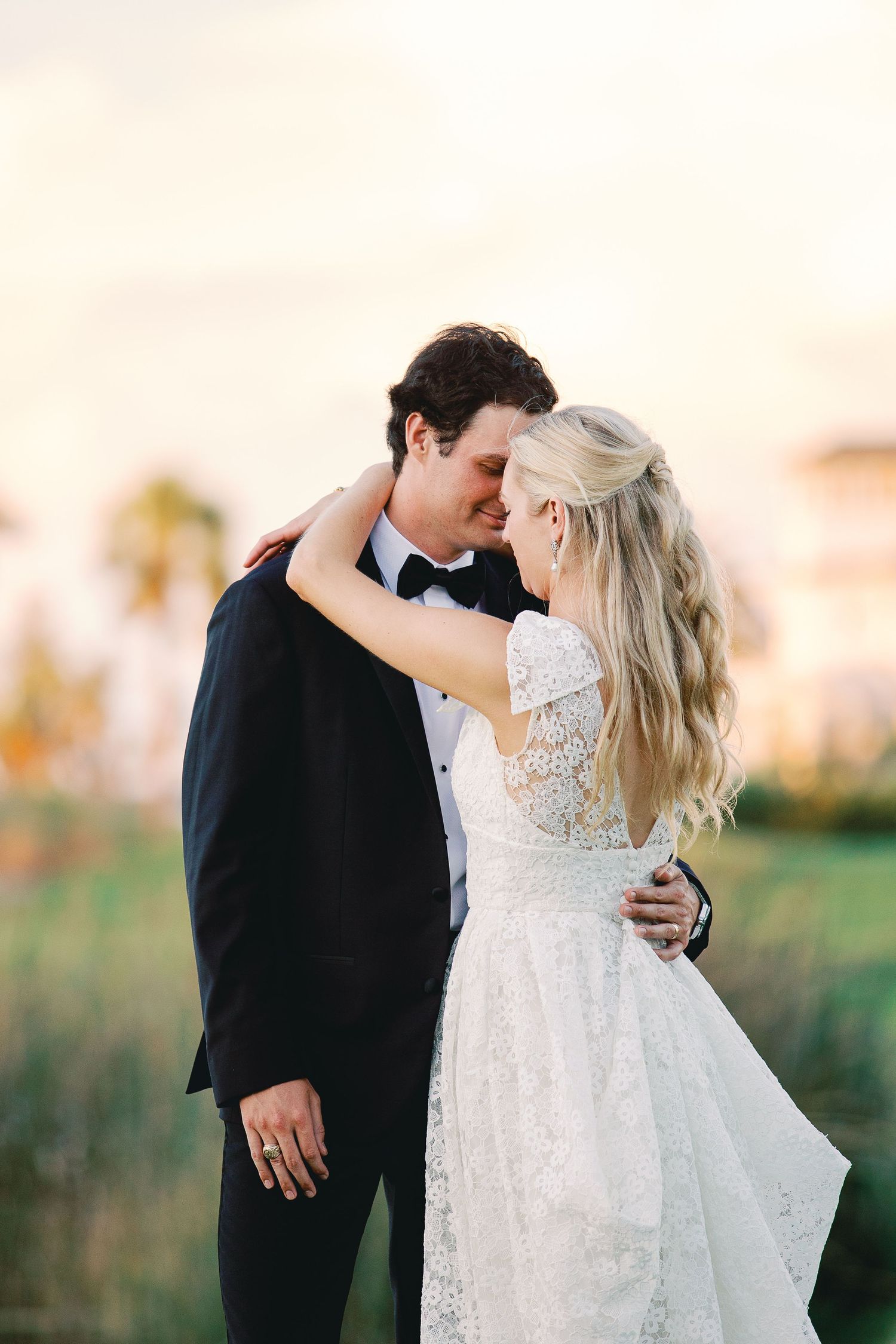 A bride and groom share a romantic moment at sunset, with her in lace gown and him in black tuxedo.