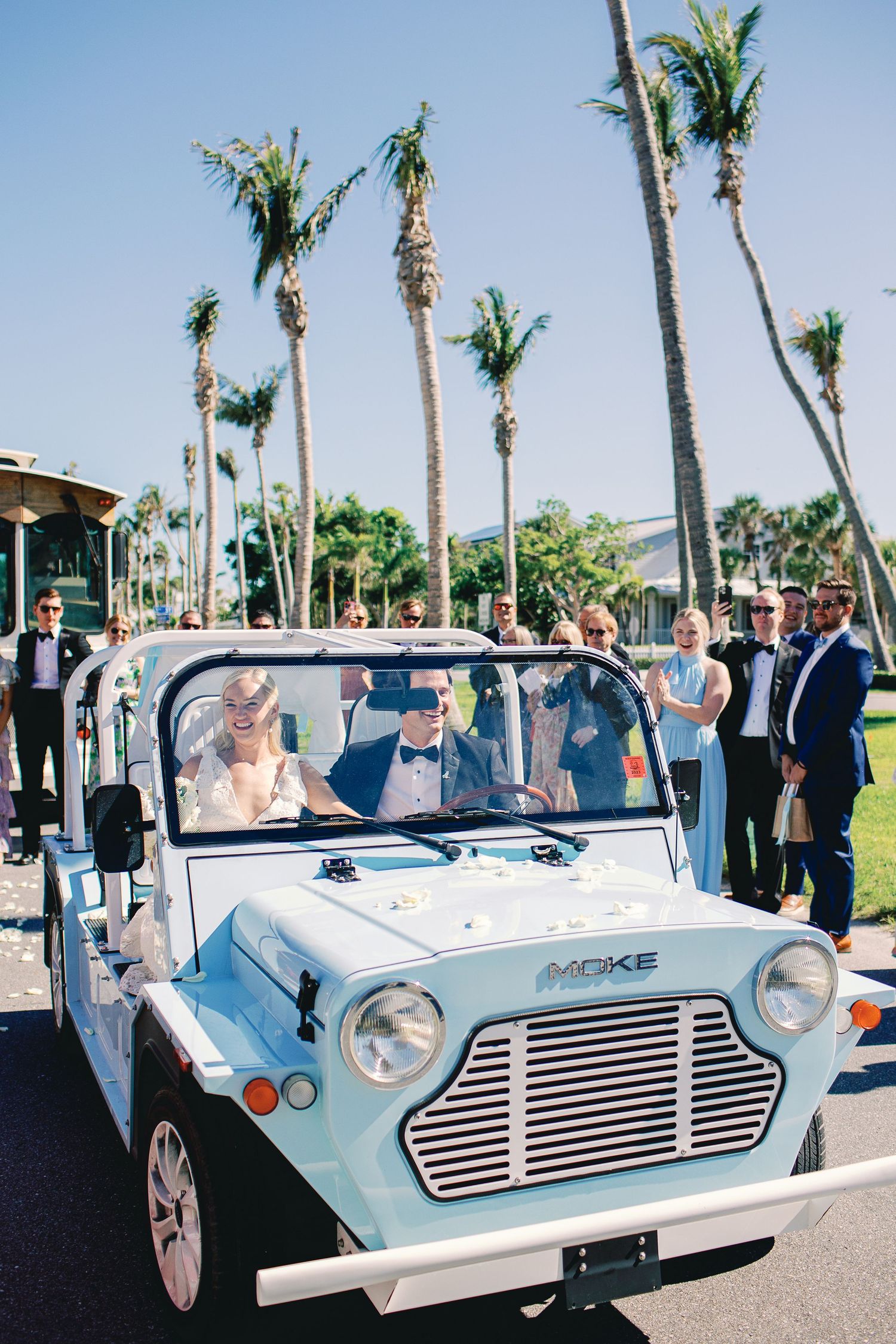 Bride and groom ride a vintage light blue Mini Moke convertible at a tropical wedding venue lined with palm trees.