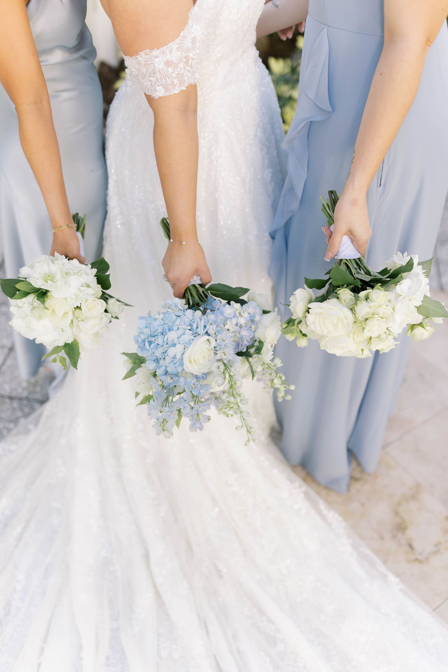 Bride holding blue hydrangea bouquet flanked by bridesmaids in dusty blue dresses with white roses.