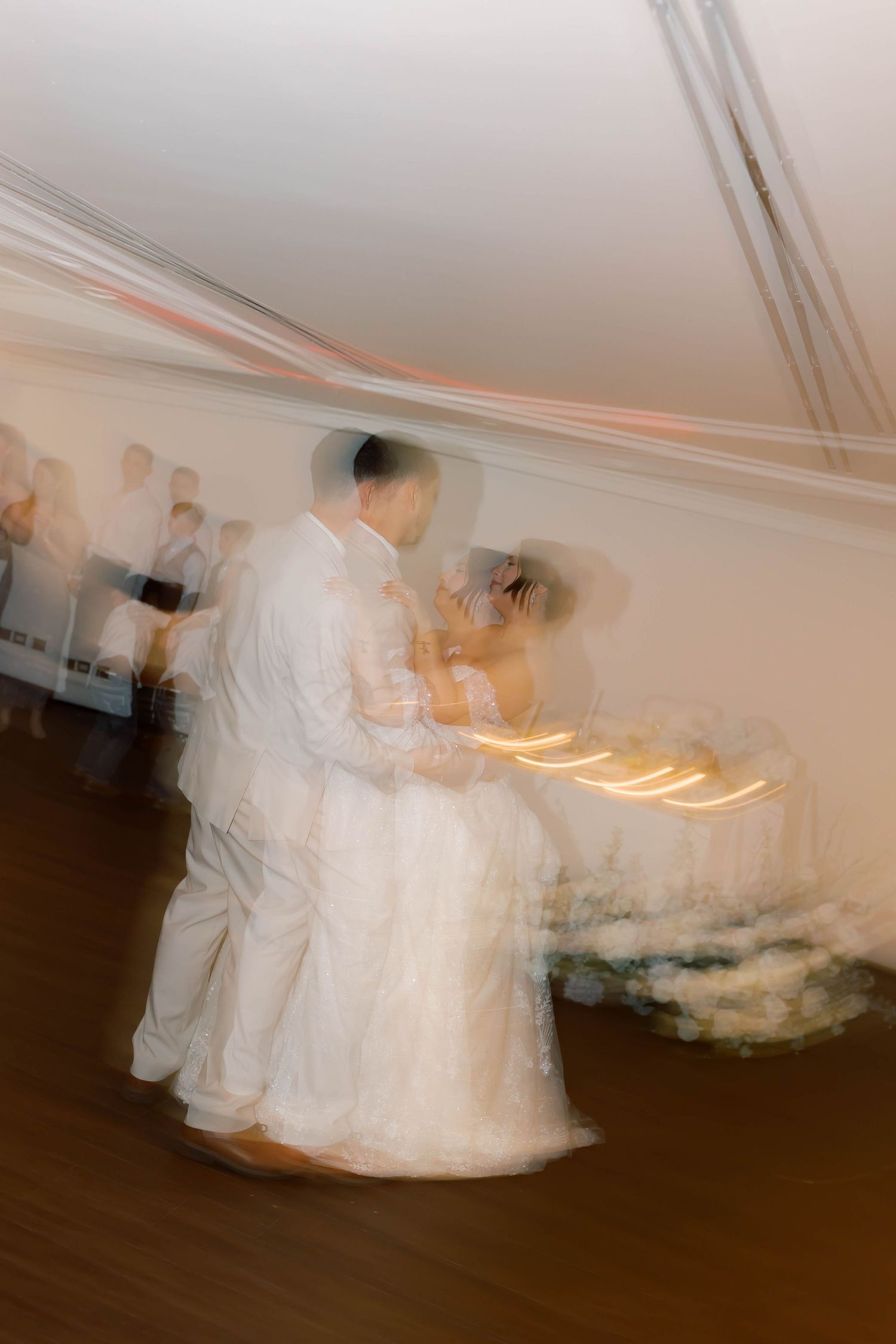 Motion blur artistic photo of bride and groom sharing their first dance under soft warm lighting at wedding reception.