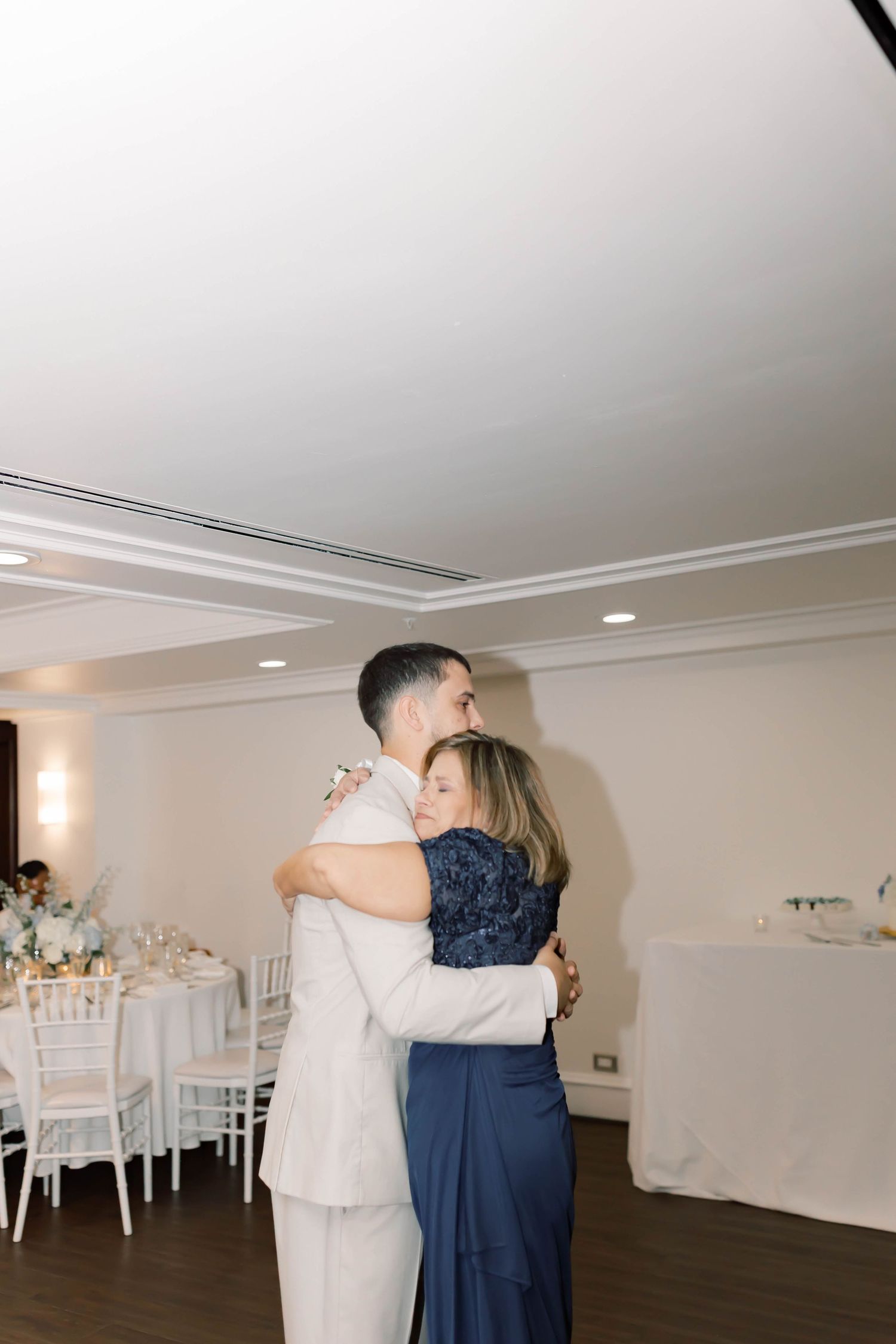 Groom in ivory suit shares emotional mother-son dance embrace in white decorated reception hall.