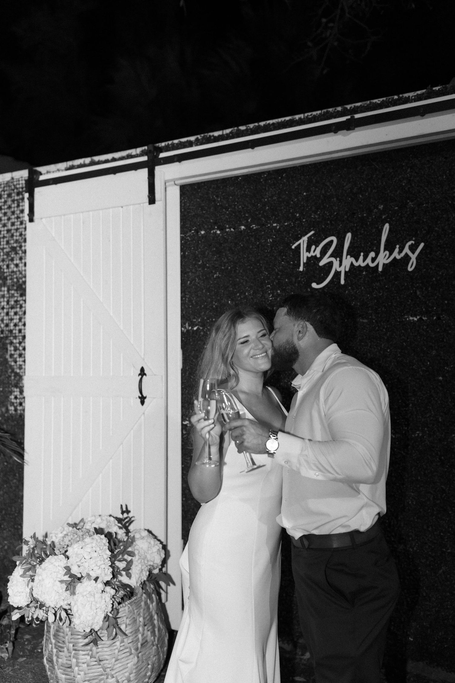 Couple sharing champagne by a white barn door with floral decorations.