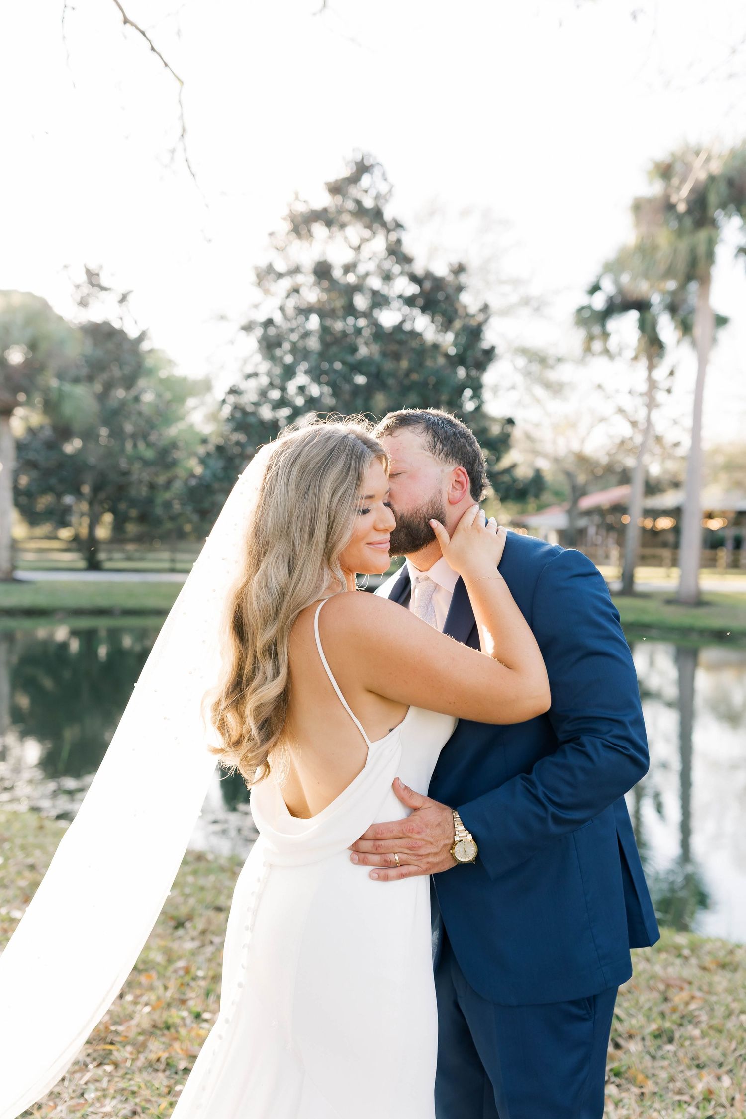 A romantic wedding portrait of a couple embracing by a lake with palm trees and greenery in the background.