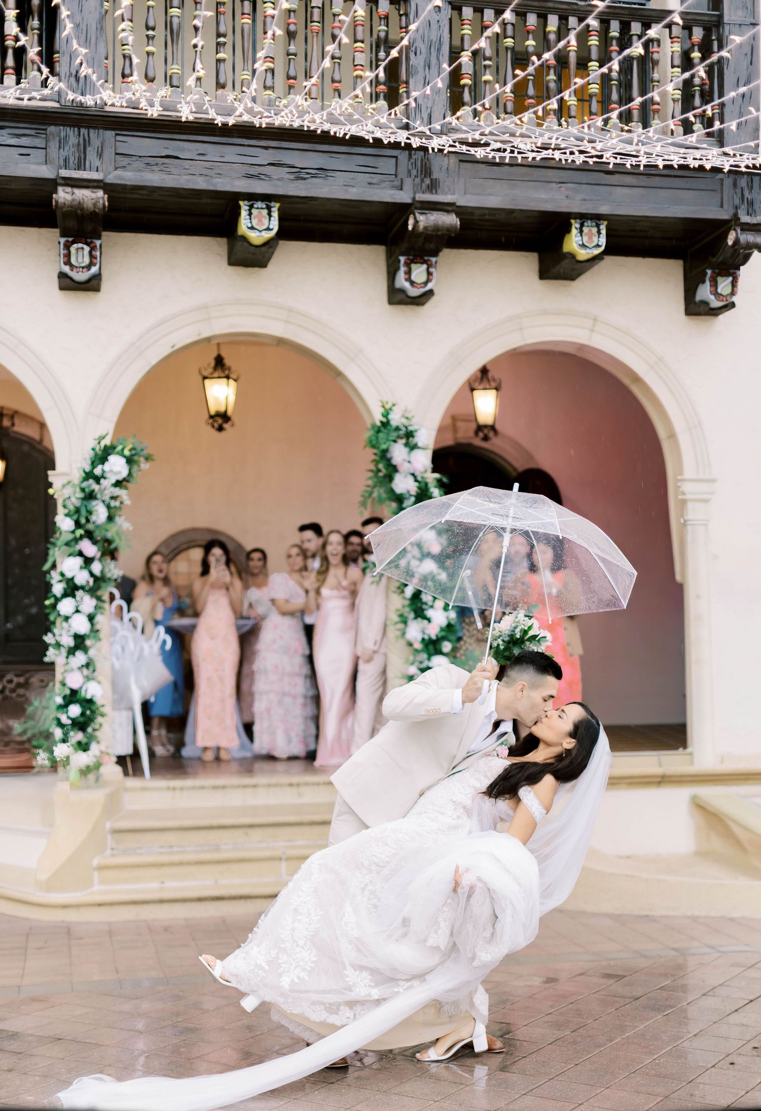 Bride and groom share a romantic dip kiss in a courtyard while holding a clear umbrella, with bridesmaids watching nearby.