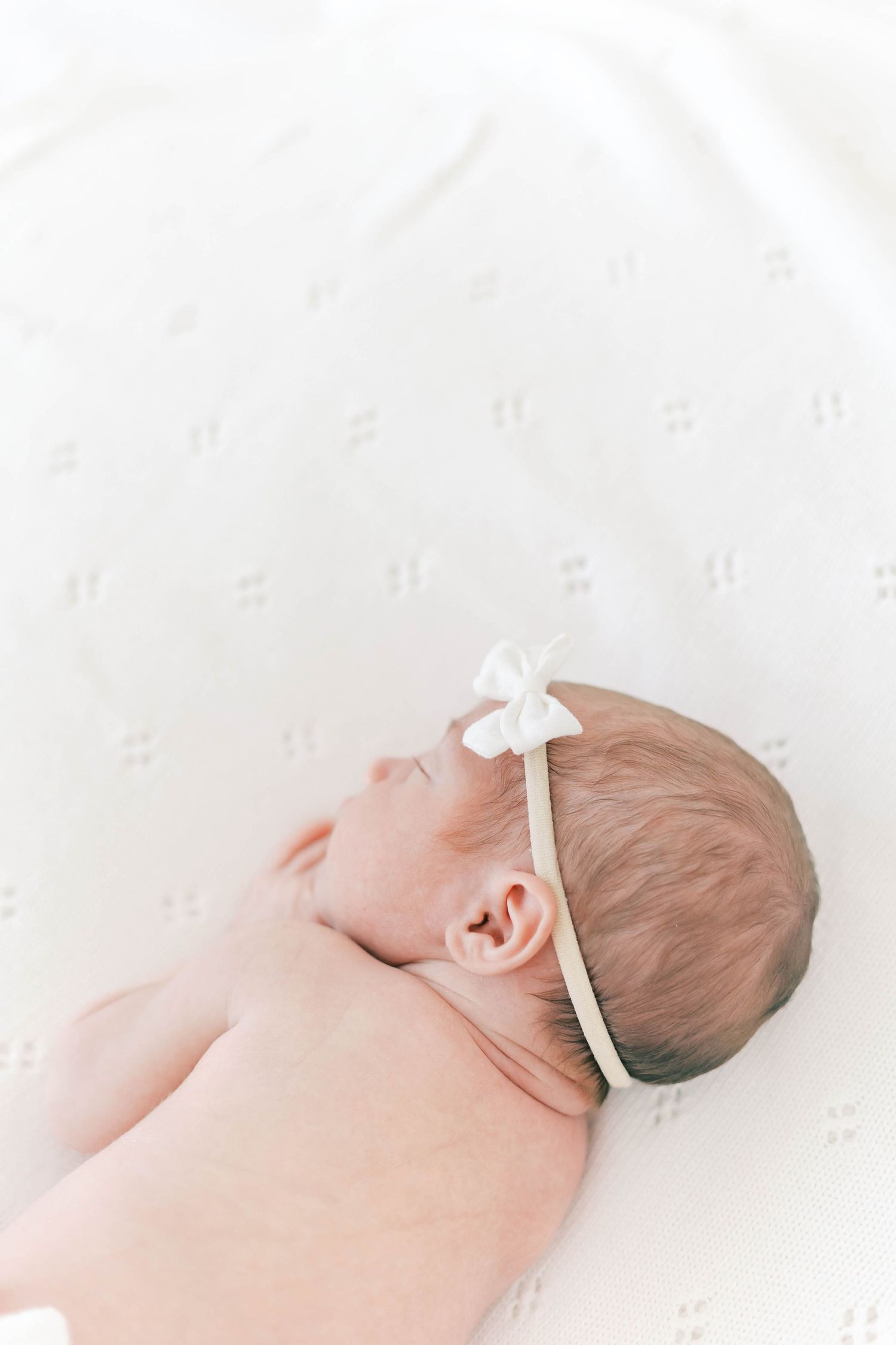 A newborn baby wearing a white bow headband rests on a white textured background.