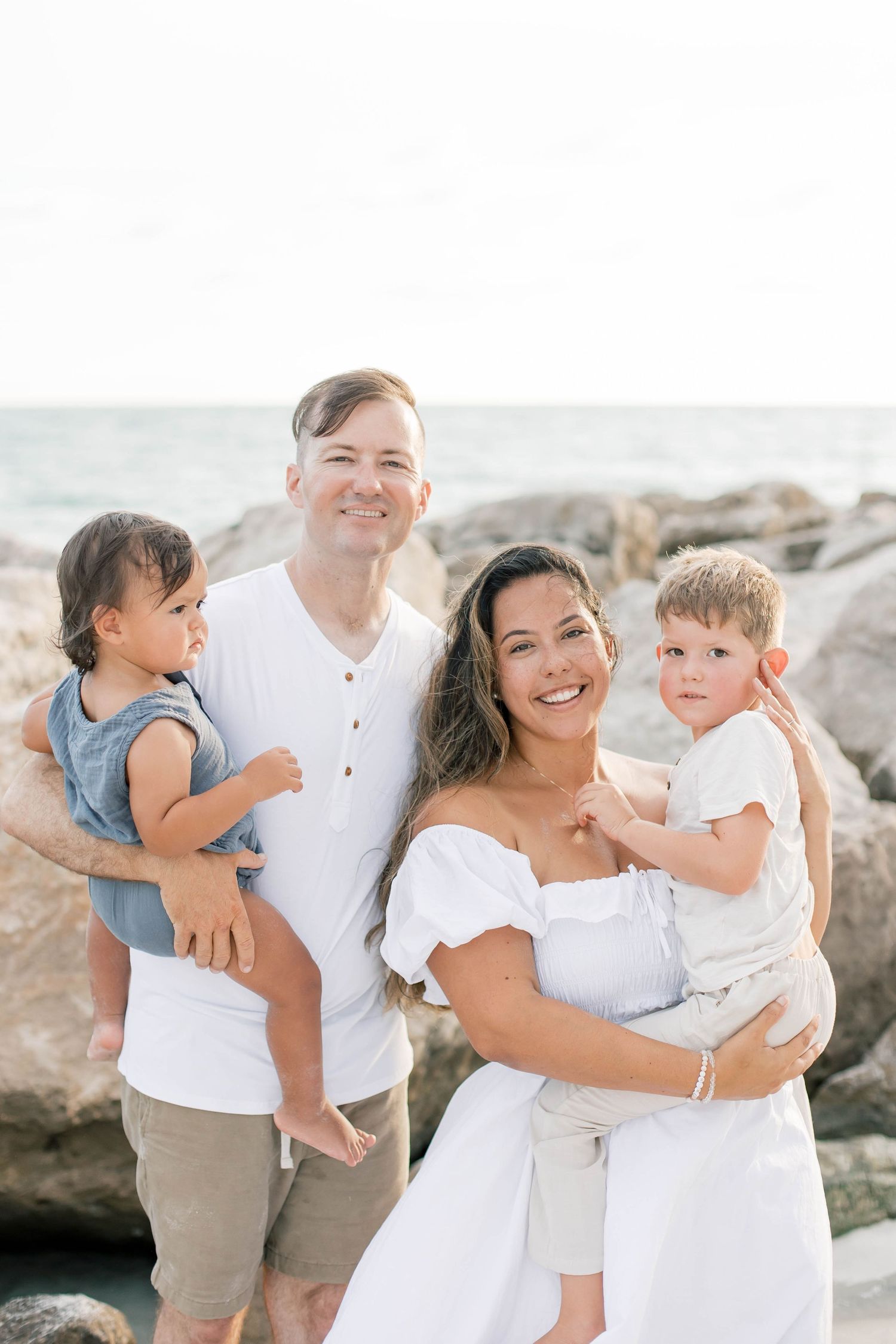 A happy family moment captured on a sunny beach with coastal rocks in the background.
