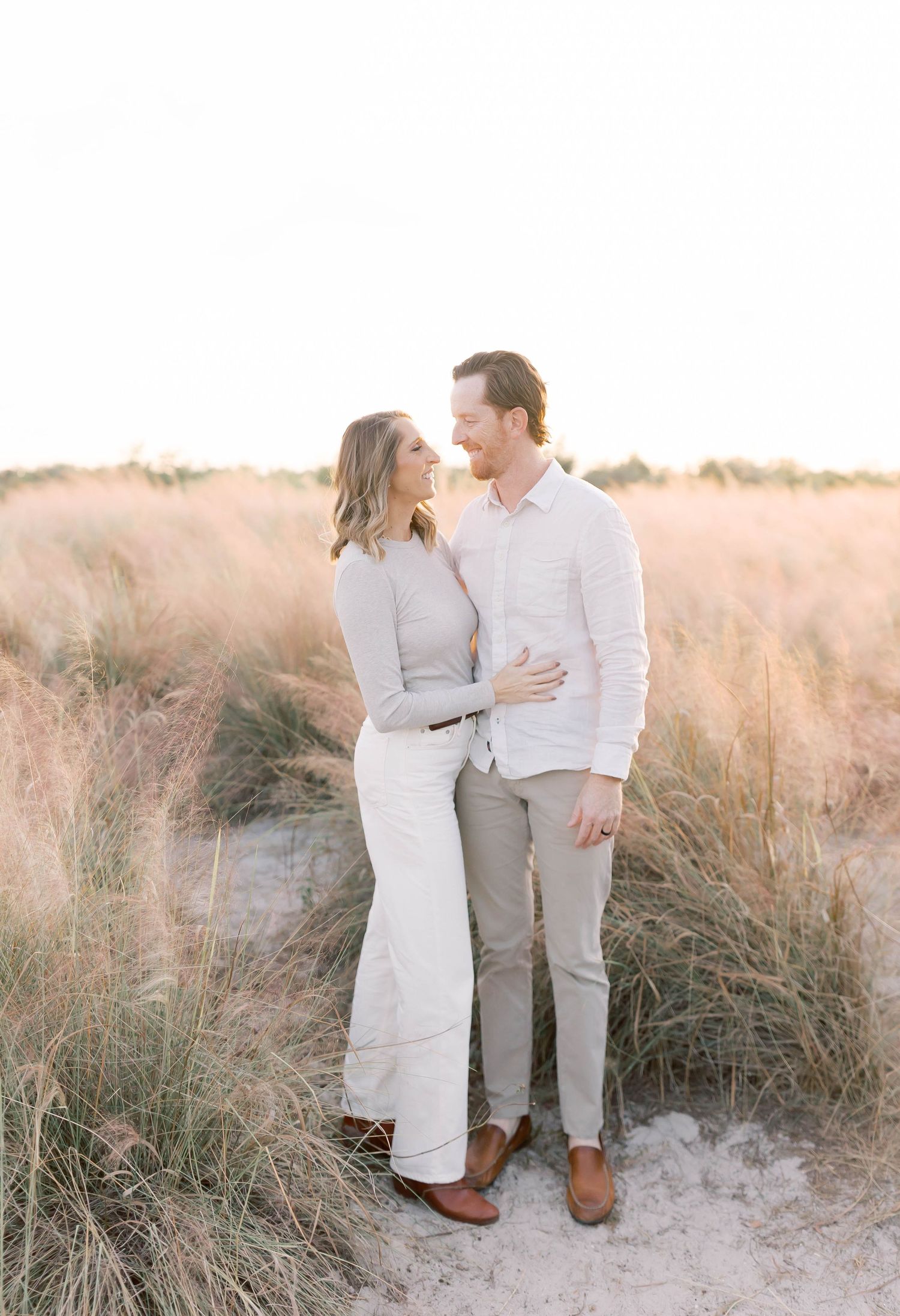 Family wearing coordinated white outfits poses together on sandy beach with tall grass backdrop.