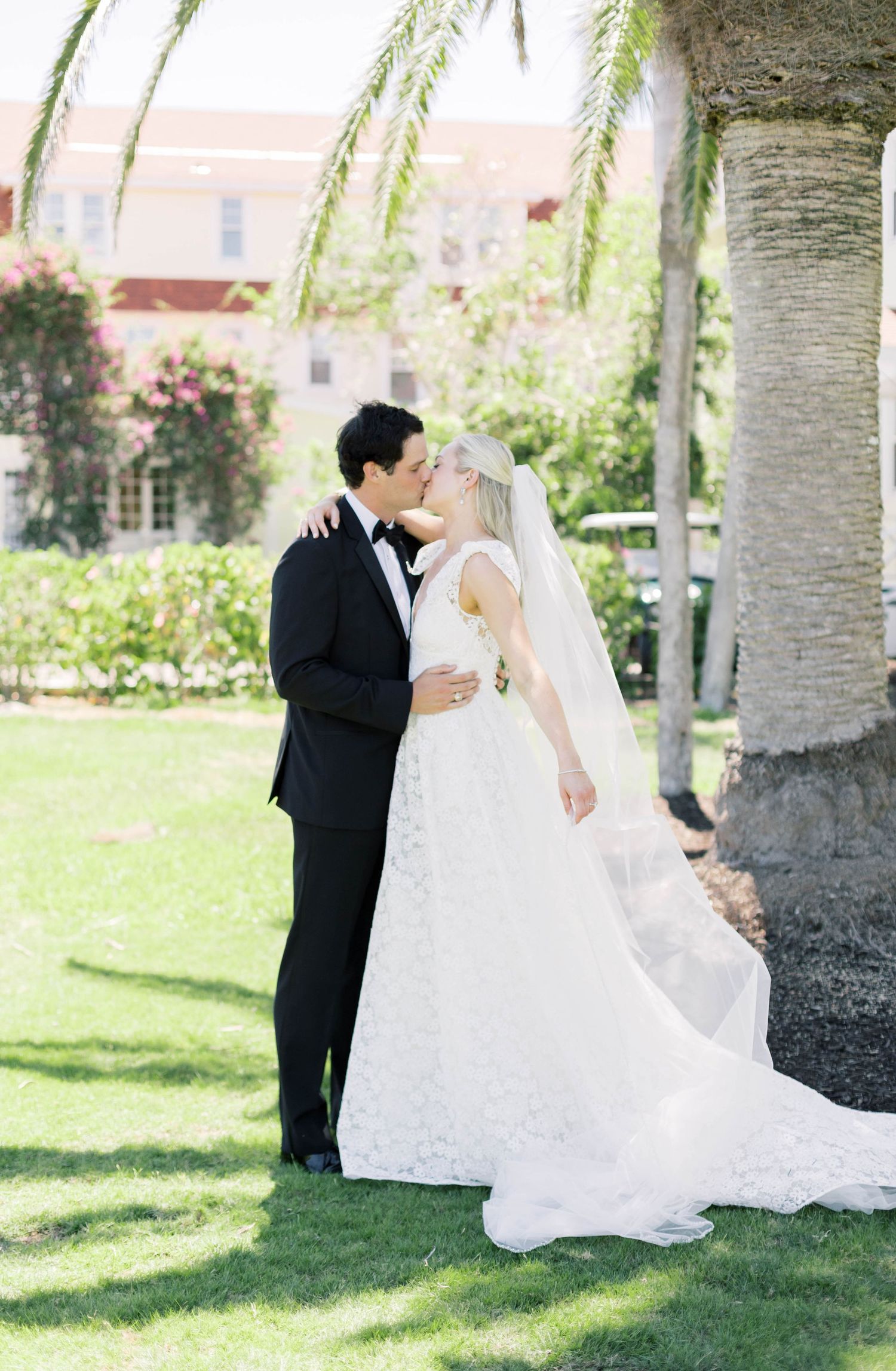 A bride and groom share a romantic kiss outdoors near a palm tree, with the bride wearing a lace gown and veil.
