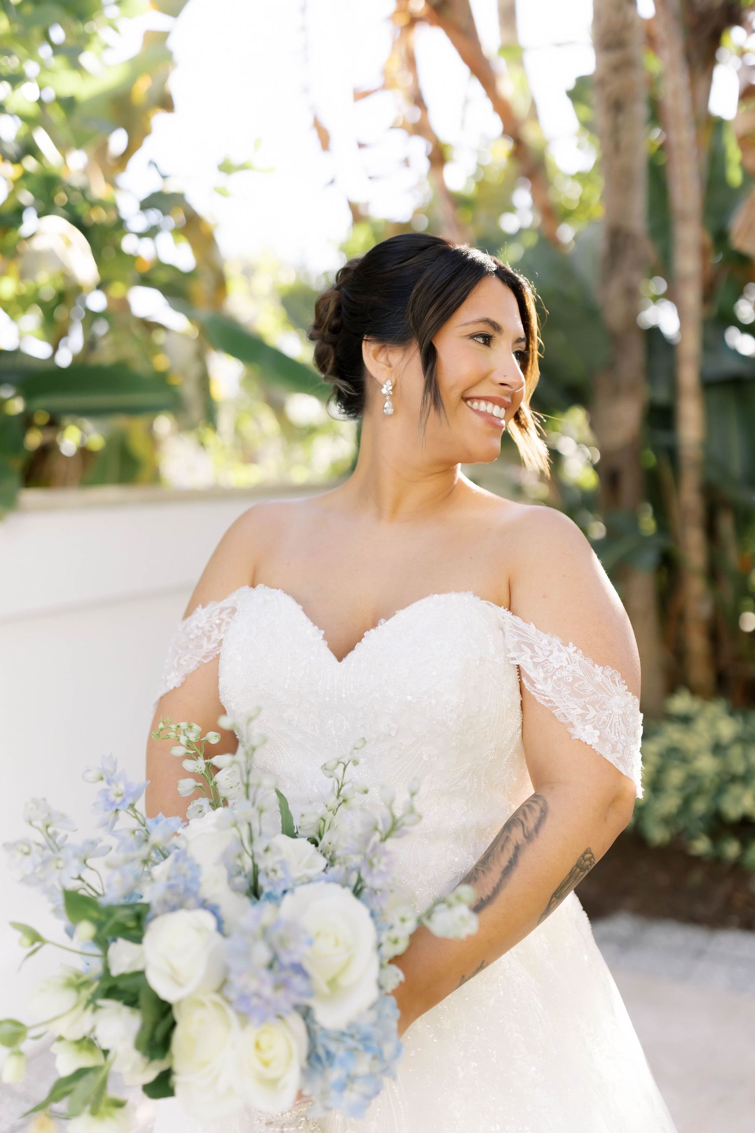 Smiling bride in off-shoulder lace gown holds blue and white bouquet of roses and delphinium in tropical garden setting.