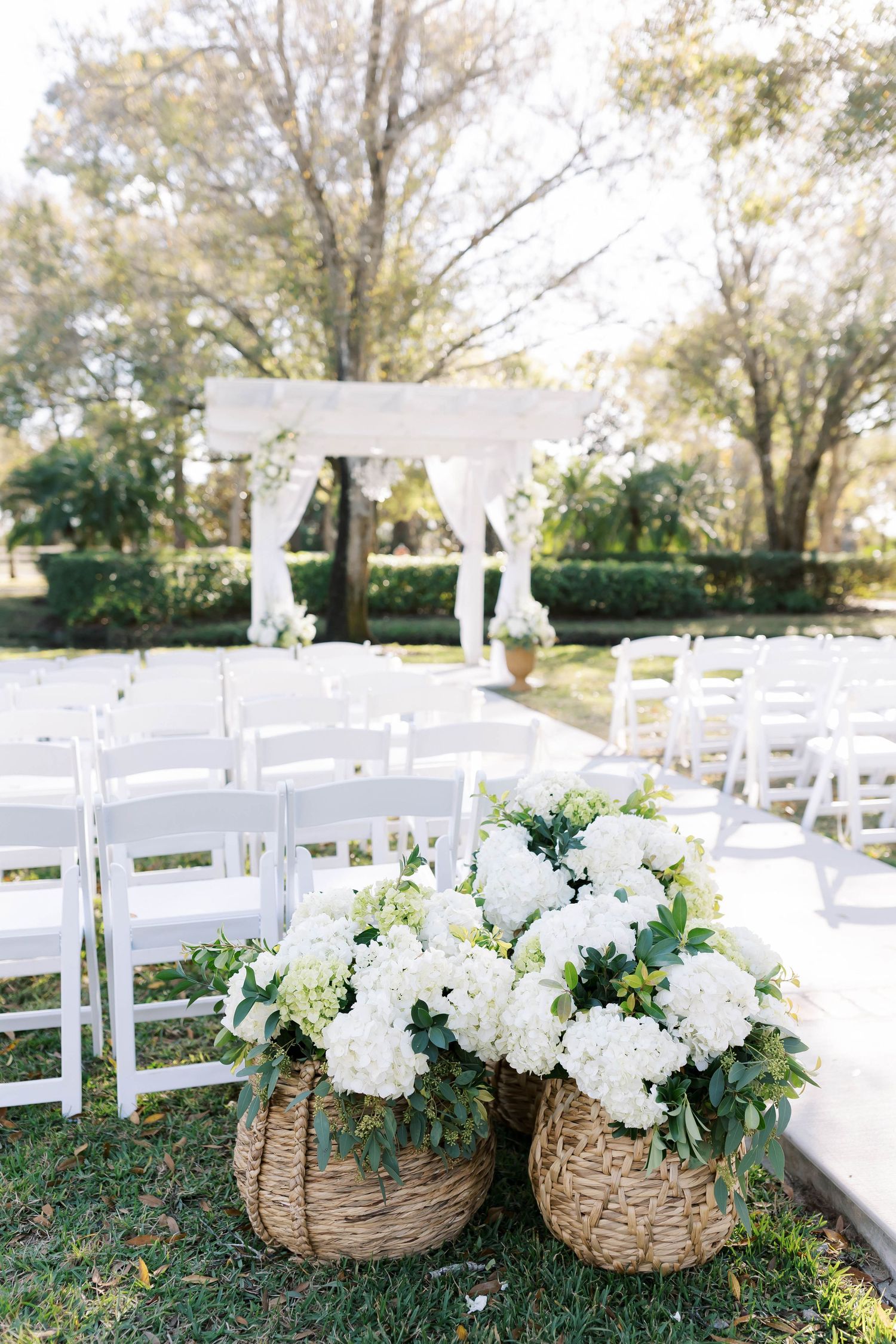 Rustic woven baskets filled with white hydrangeas decorate a white wedding ceremony aisle.