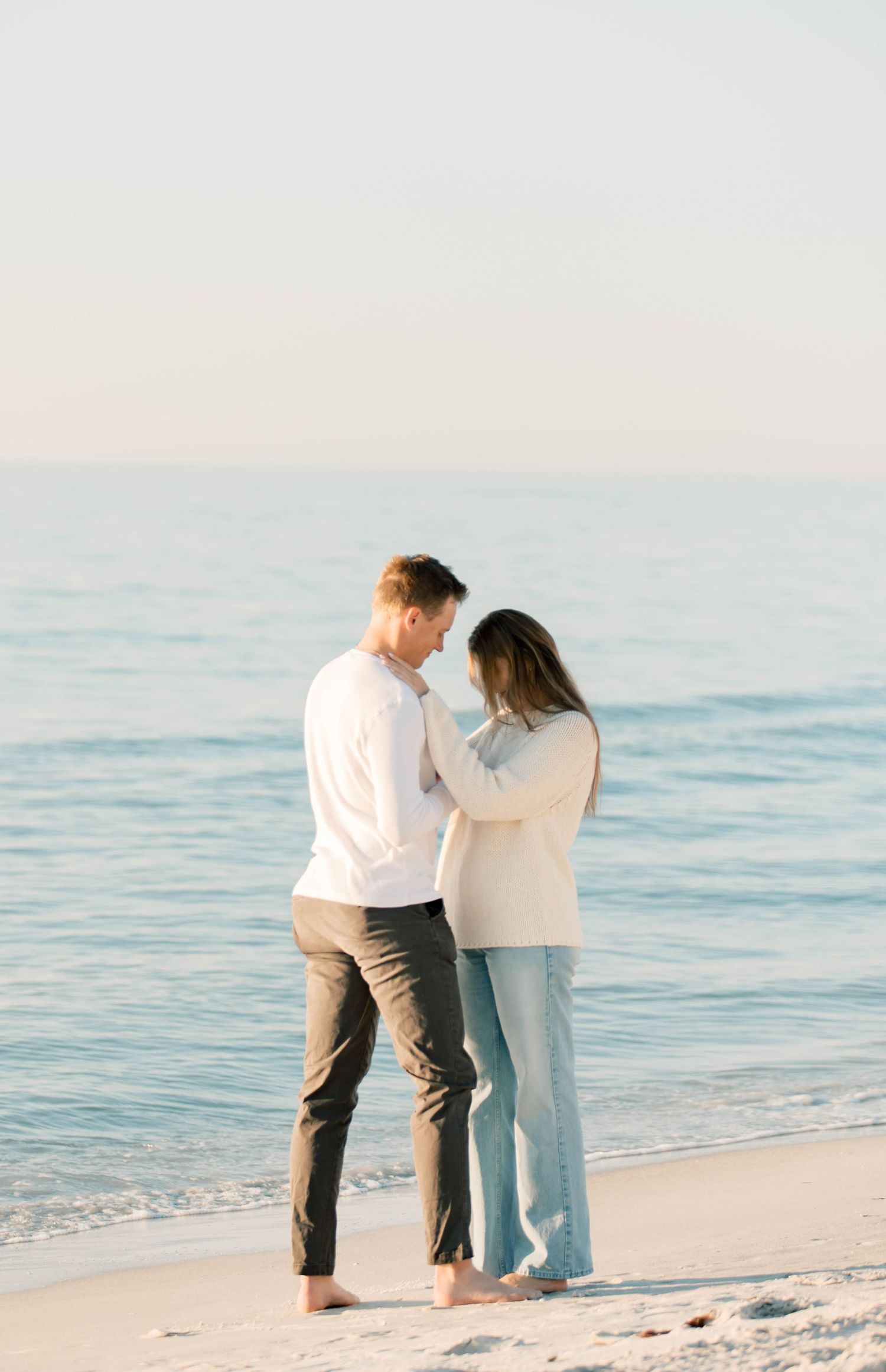 Two people in white and dark clothing walk together along a peaceful beach at sunset.