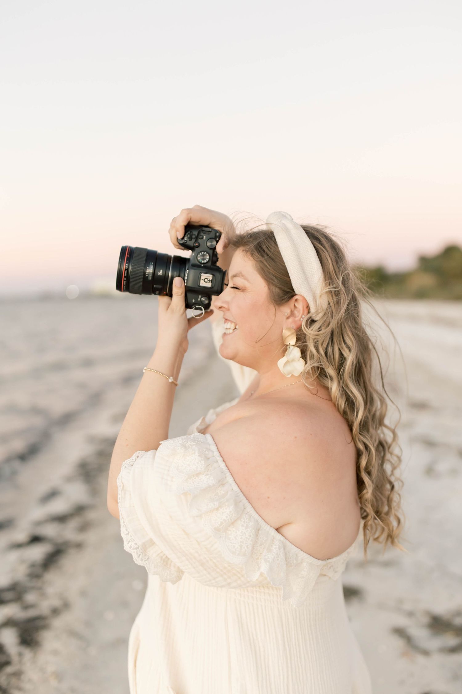 Smiling woman in white off-shoulder dress holding DSLR camera on beach at sunset with wavy hair and gold earrings.
