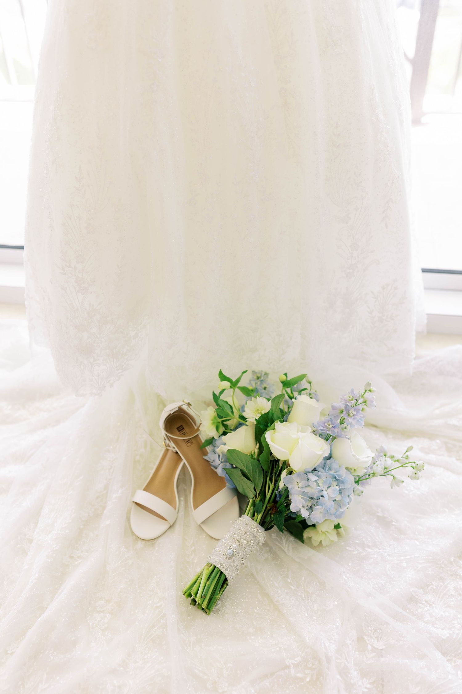 White lace wedding dress with bridal bouquet of blue hydrangeas and white roses beside strappy white heels on floor.