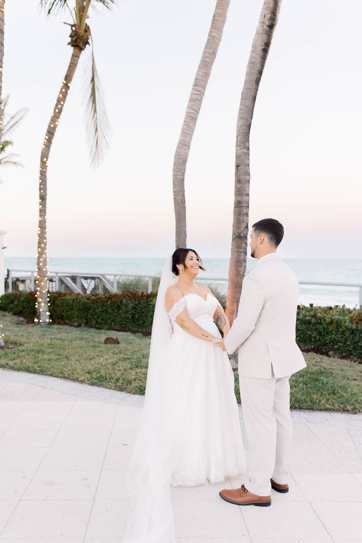 Bride and groom hold hands facing each other near palm trees with ocean views at a beachside wedding.