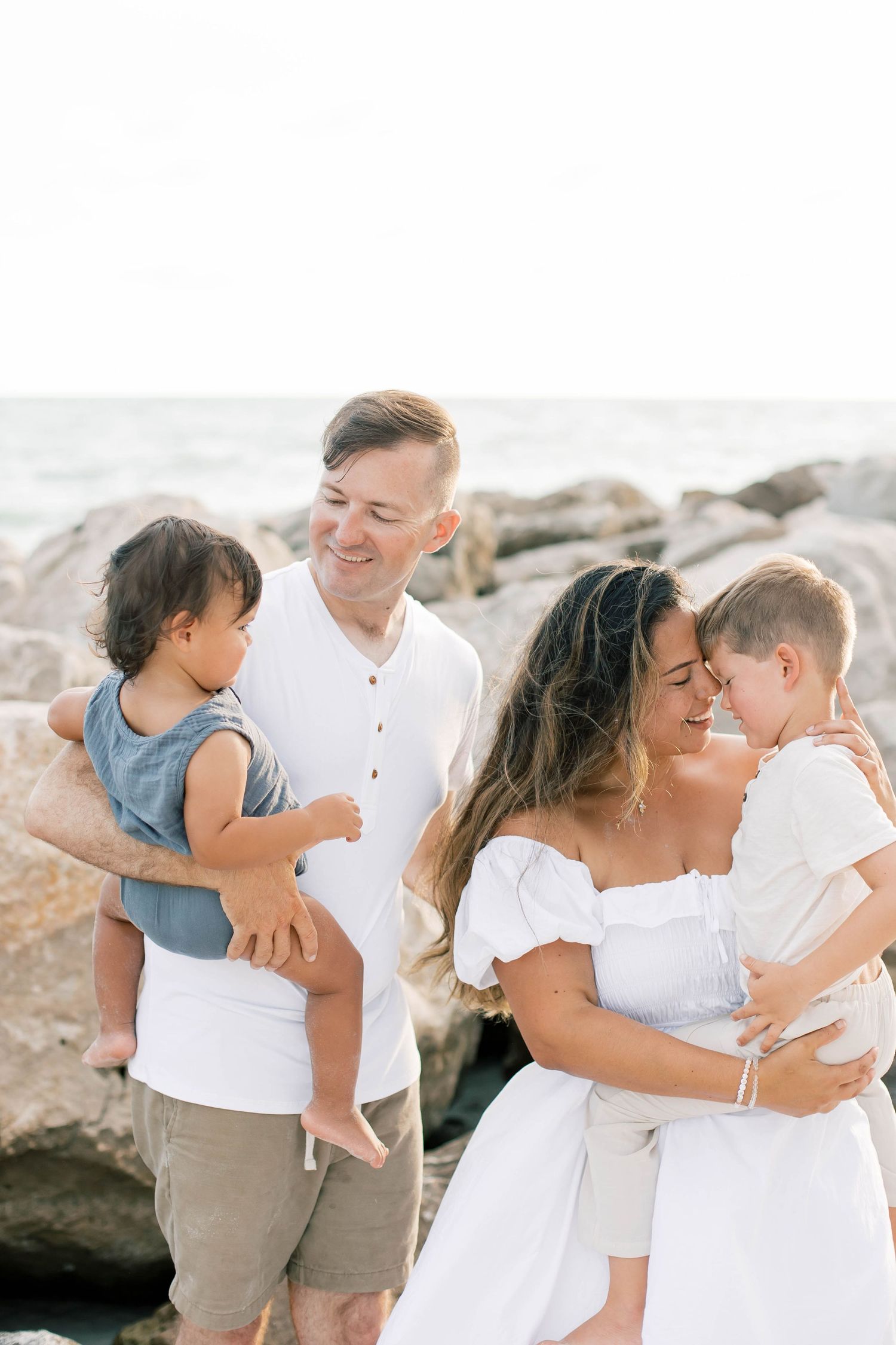 A family shares tender moments together on a beach with rocky formations in the background.