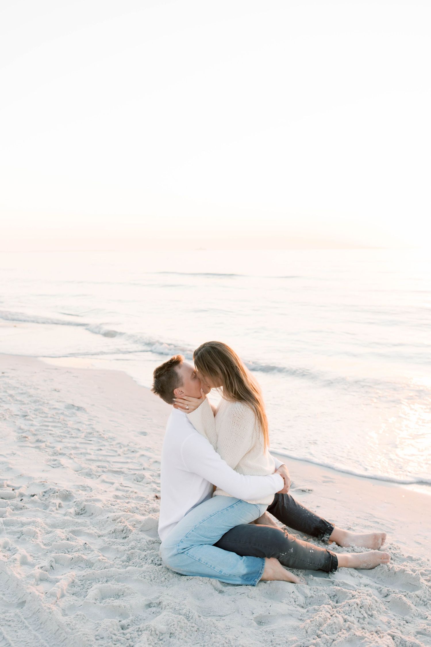Couple embracing on sandy beach during sunset wearing matching white sweaters.