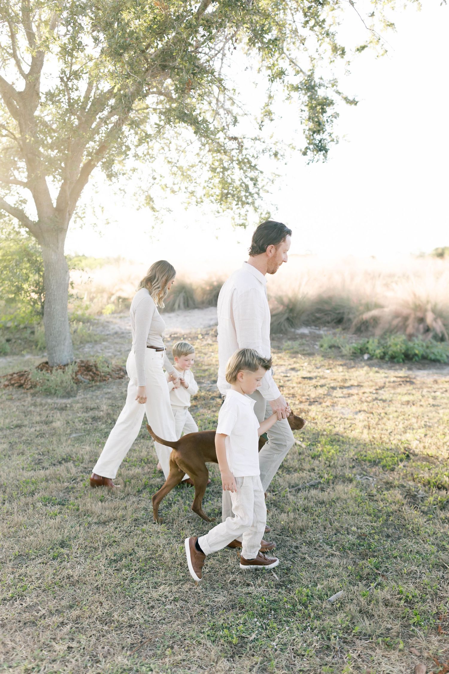 Family in white outfits plays together under trees with their brown dog.