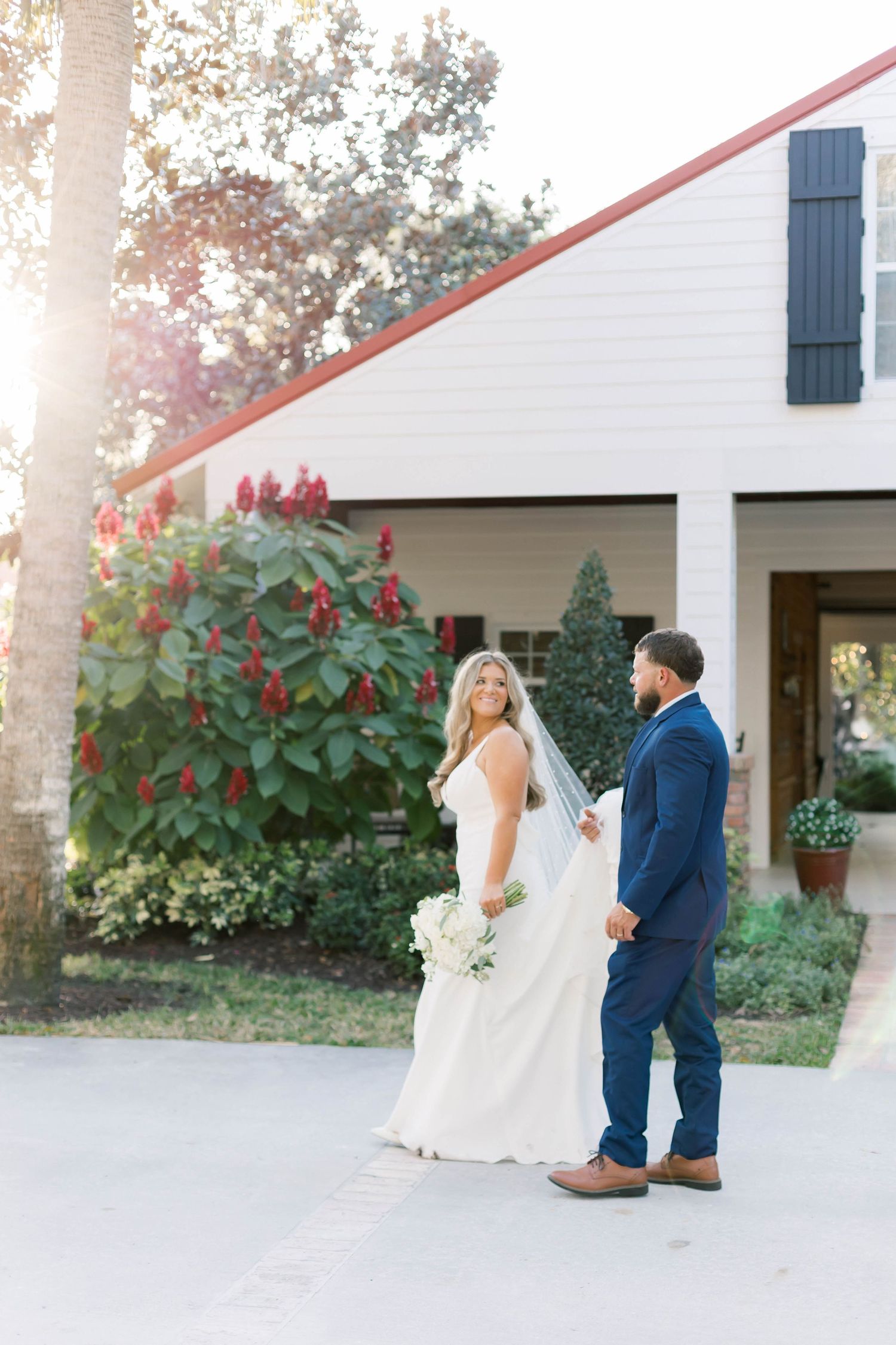 Wedding couple standing outside a white farmhouse with red flowers and greenery.