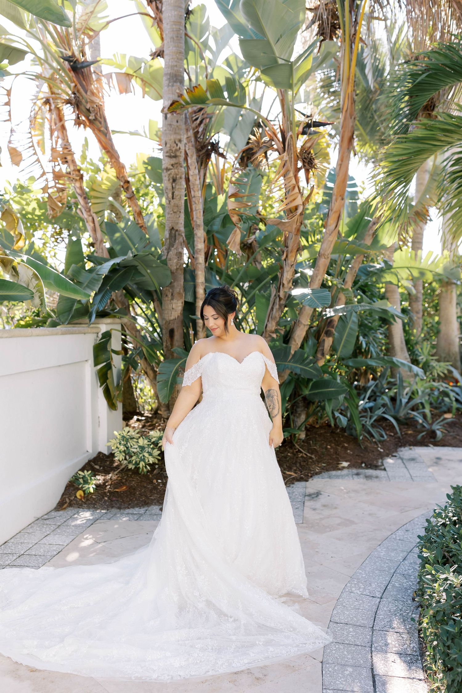 Bride in elegant off-shoulder lace gown with long train poses among tropical palm trees.