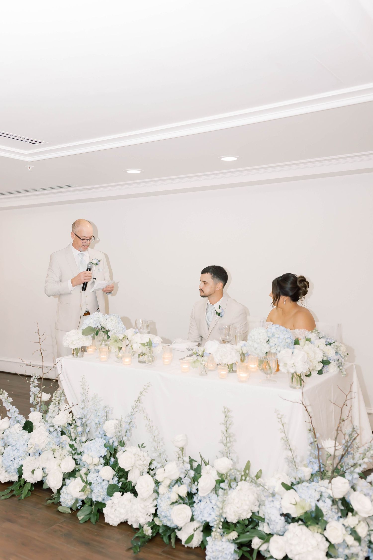 Wedding sweetheart table adorned with white and blue hydrangea floral arrangements during reception speech.