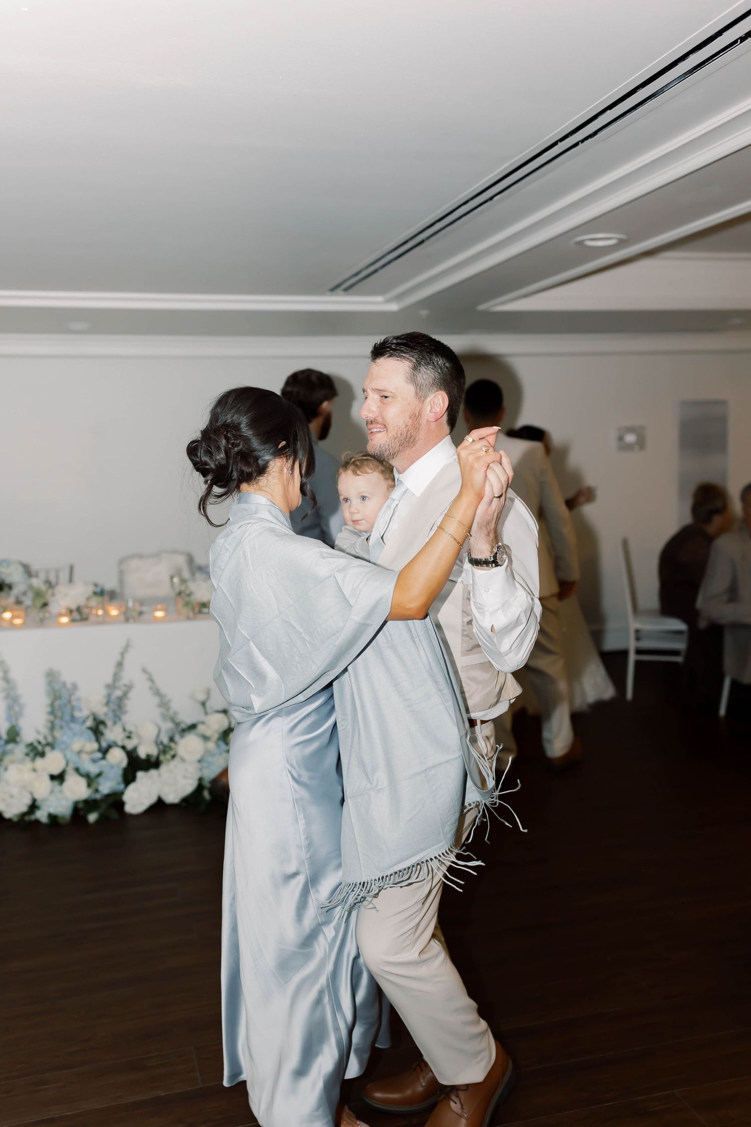 Bride dances with an older gentleman holding a child on a white-decorated wedding reception dance floor.