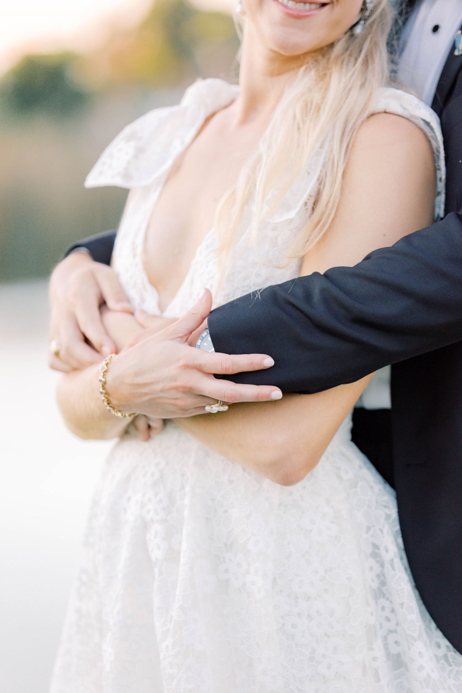 Bride in lace gown with groom embracing her from behind, showcasing wedding rings and gold bracelet.