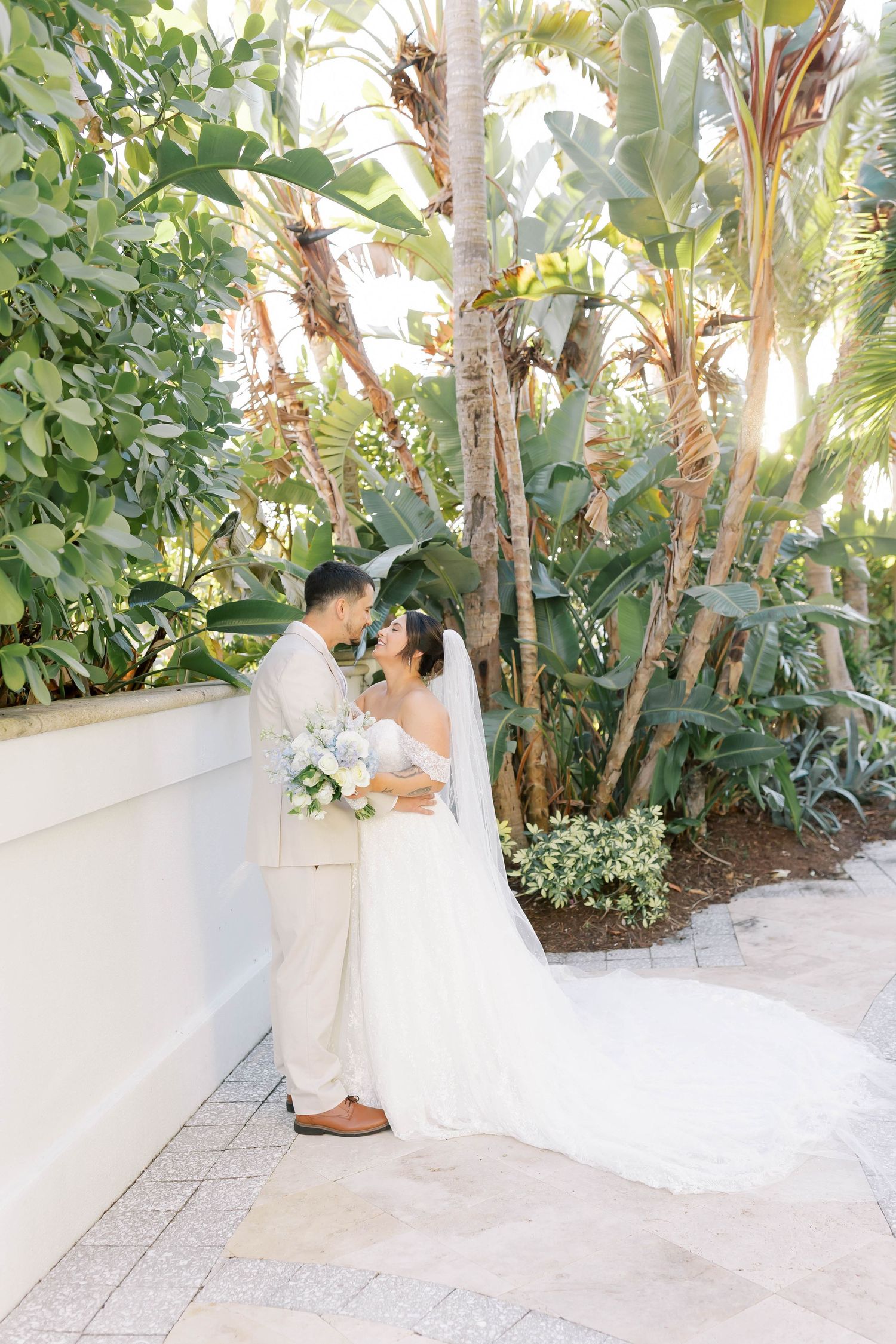 Bride and groom share a romantic moment surrounded by lush tropical greenery, bride in white gown with veil holding bouquet.