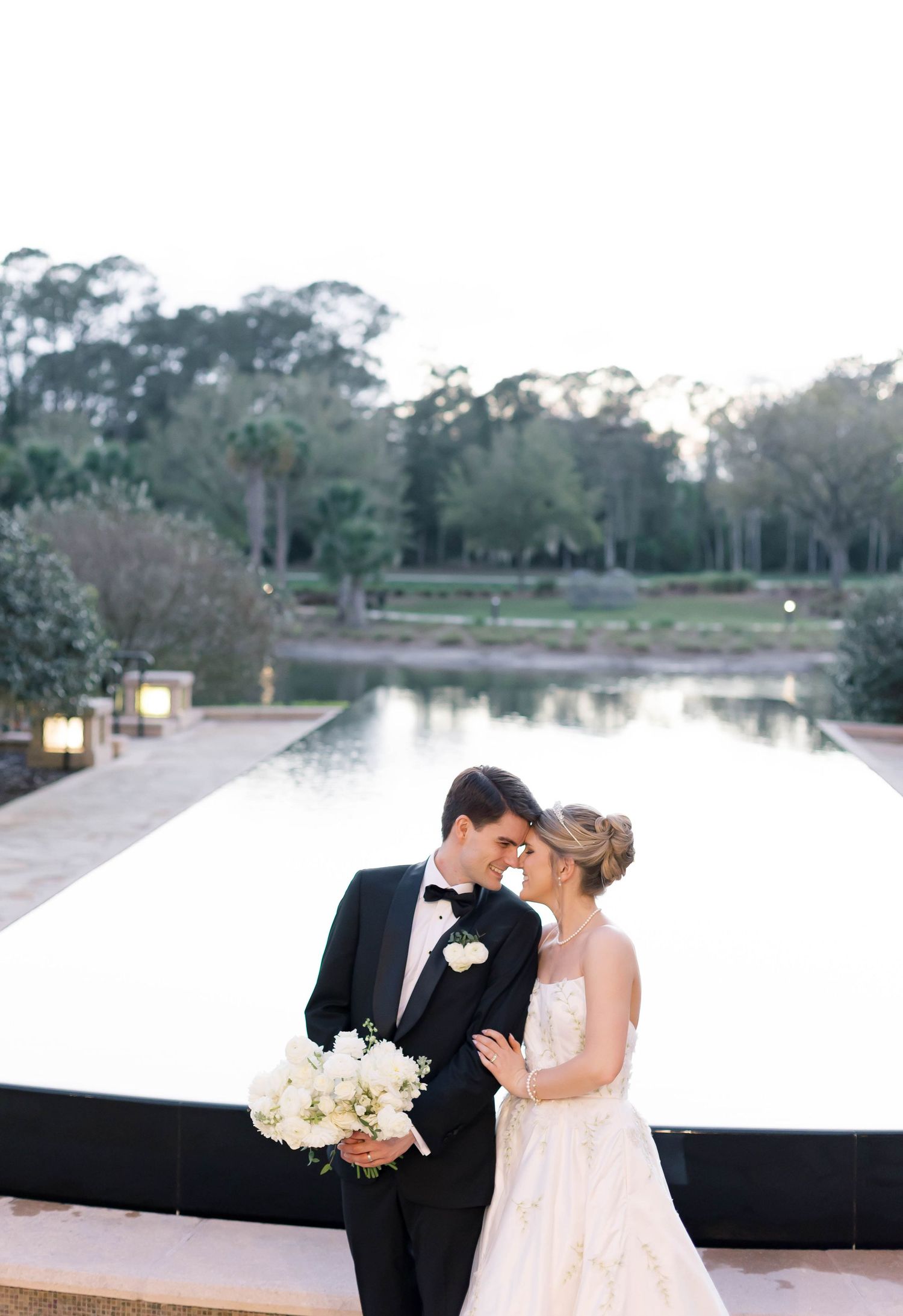 Bride and groom share romantic moment by reflective pool, she holds white bouquet, he wears black tuxedo, lush trees behind.