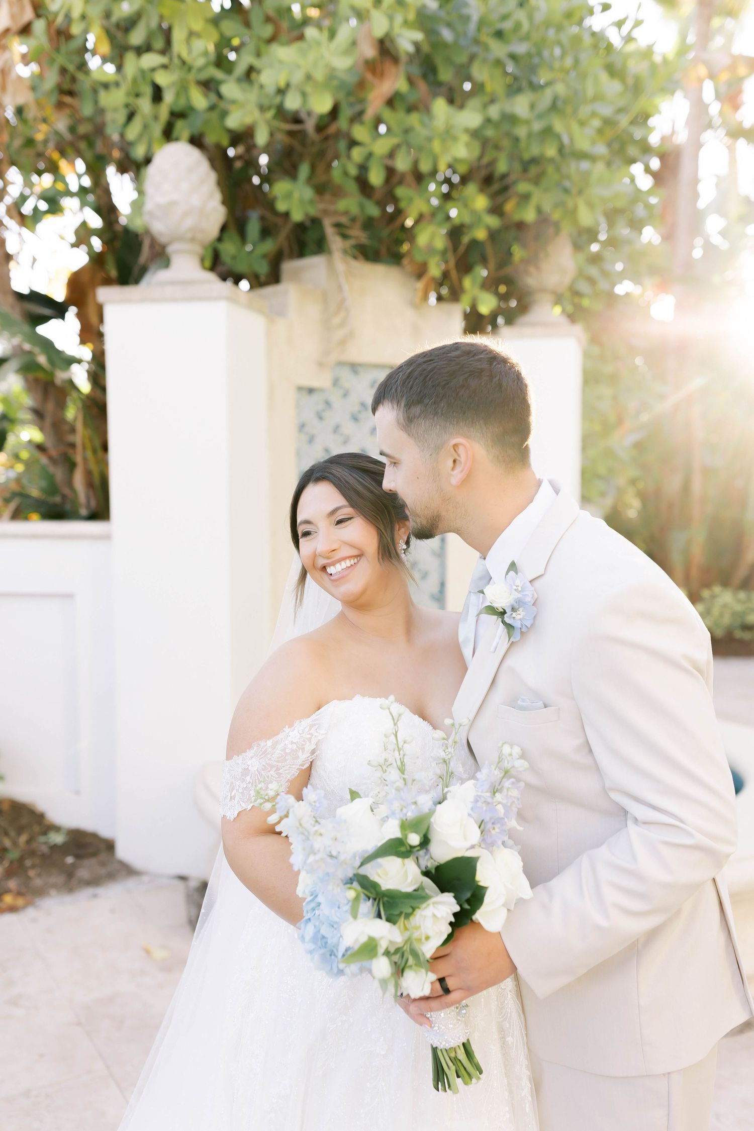 Smiling bride in off-shoulder lace gown holds blue and white bouquet as groom in tan suit whispers in her ear.