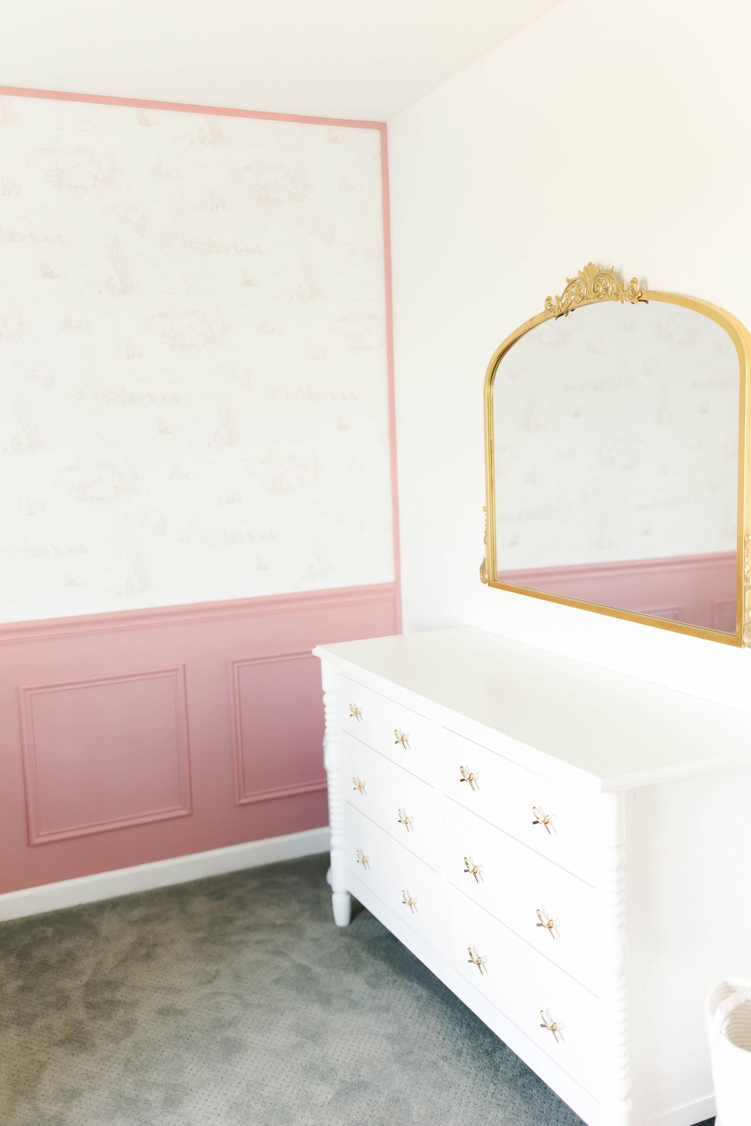 White dresser with gold mirror and pink accent wall in a minimalist nursery design.