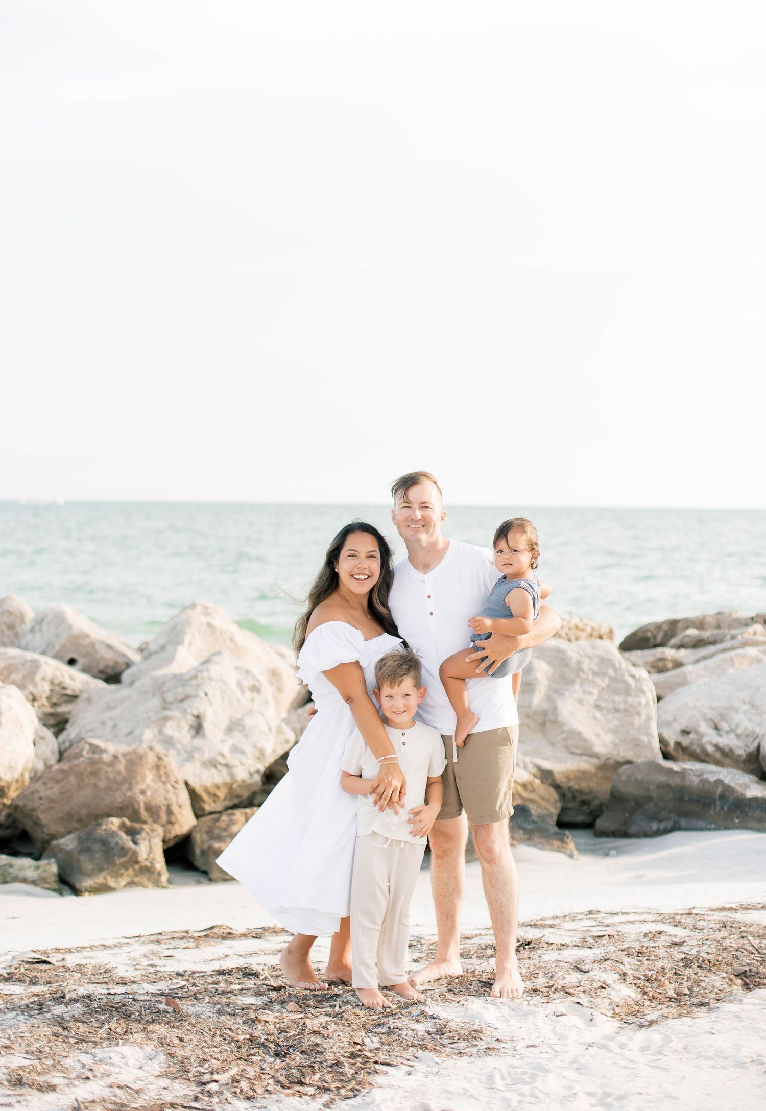 Family relaxes together on rocky beach wearing coordinated white outfits.